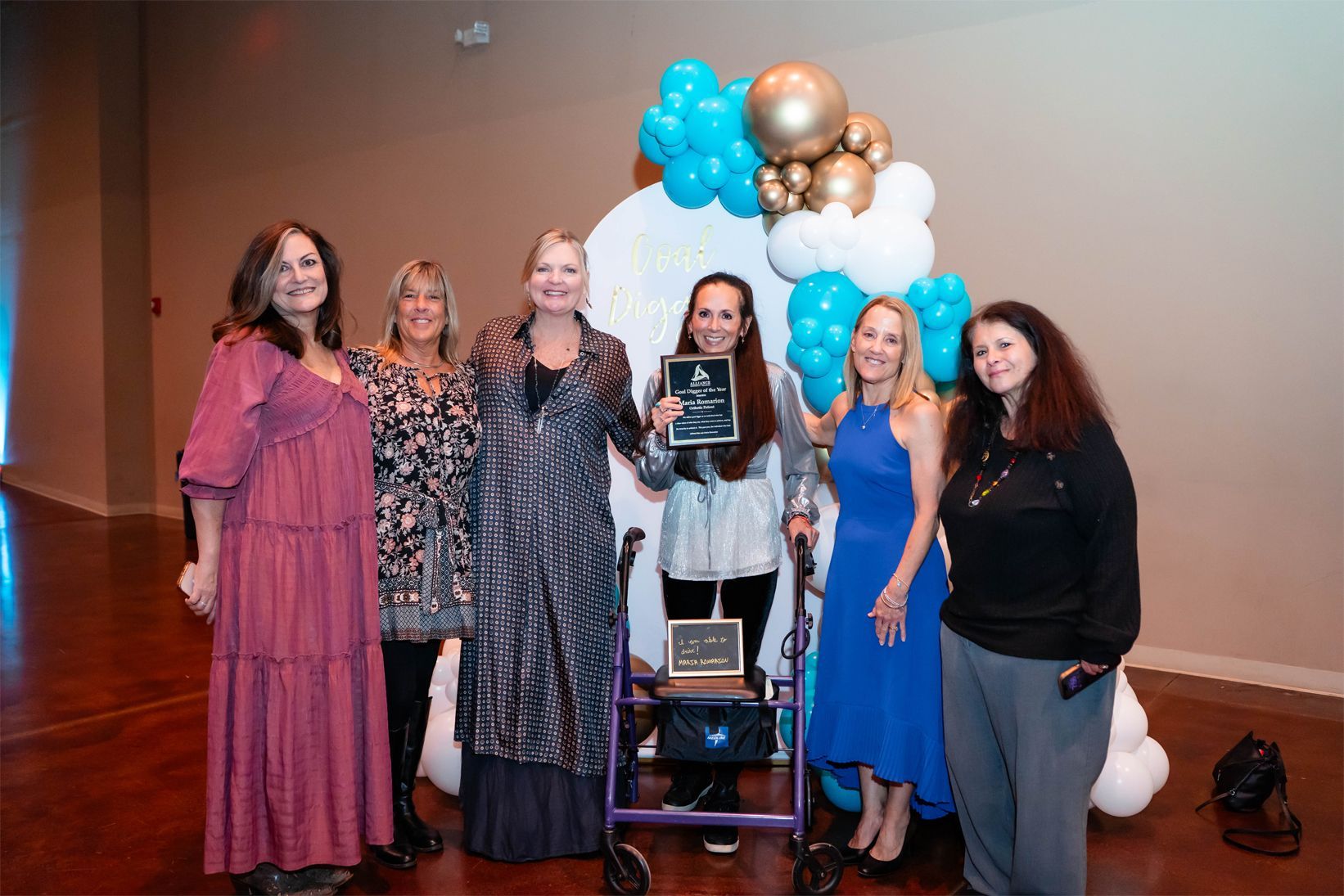 Six people stand in a line, with the person in the middle holding an award next to a walker, before a balloon display.