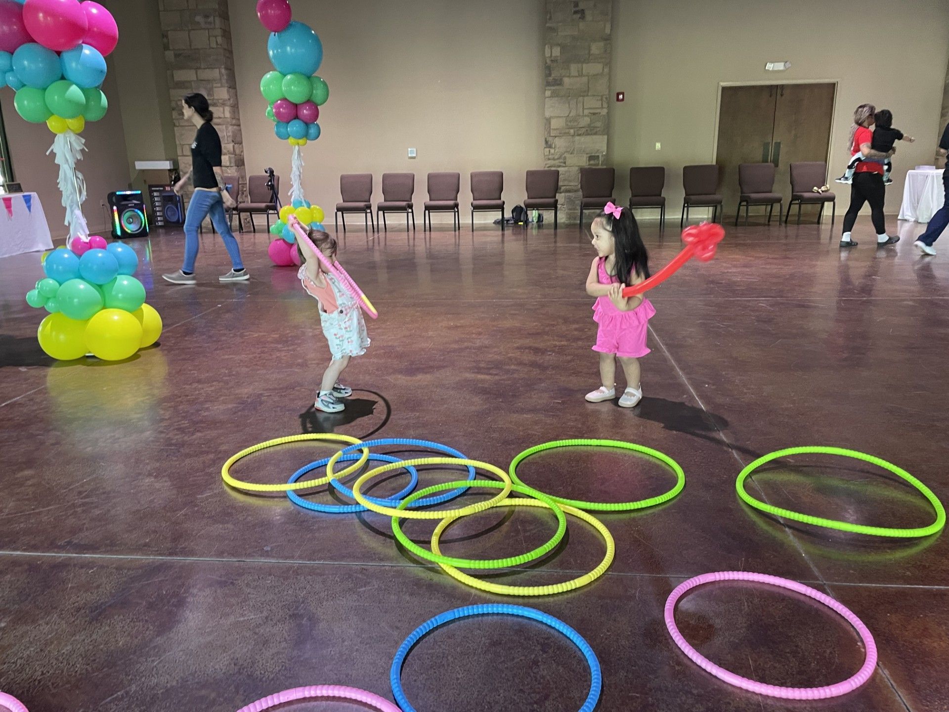 Children play with hula hoops in a hall decorated with balloons and chairs along the wall.