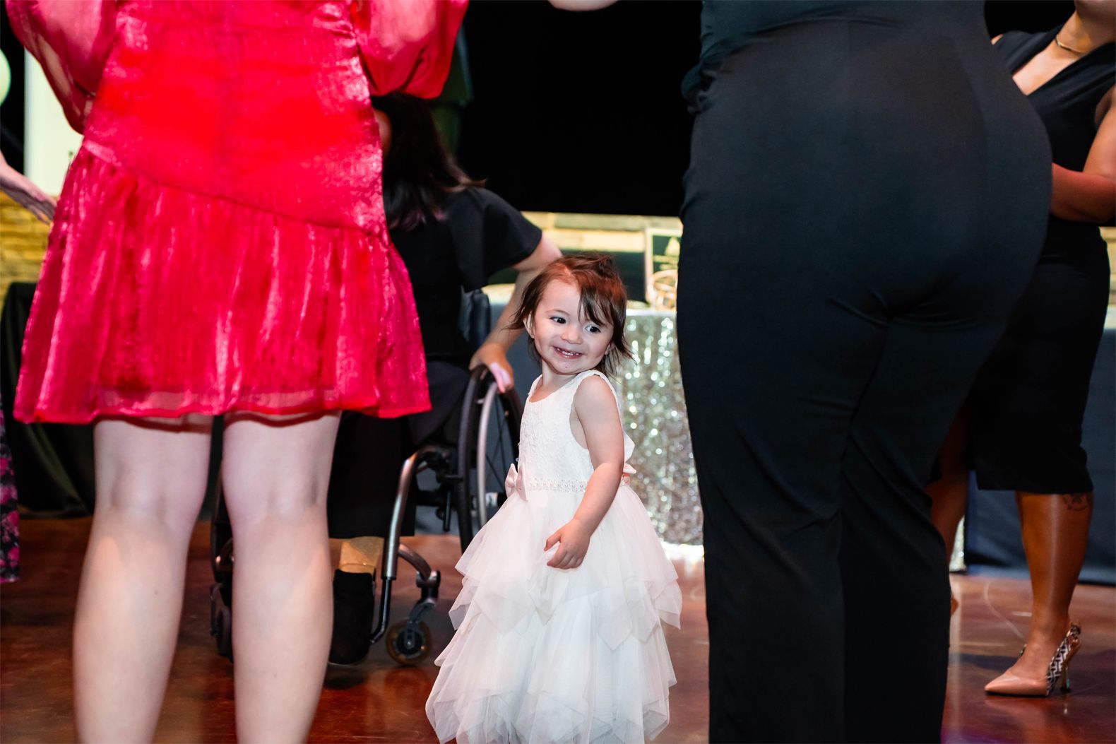 A young child in a white ruffled dress stands smiling between two people in an indoor, event-style setting.