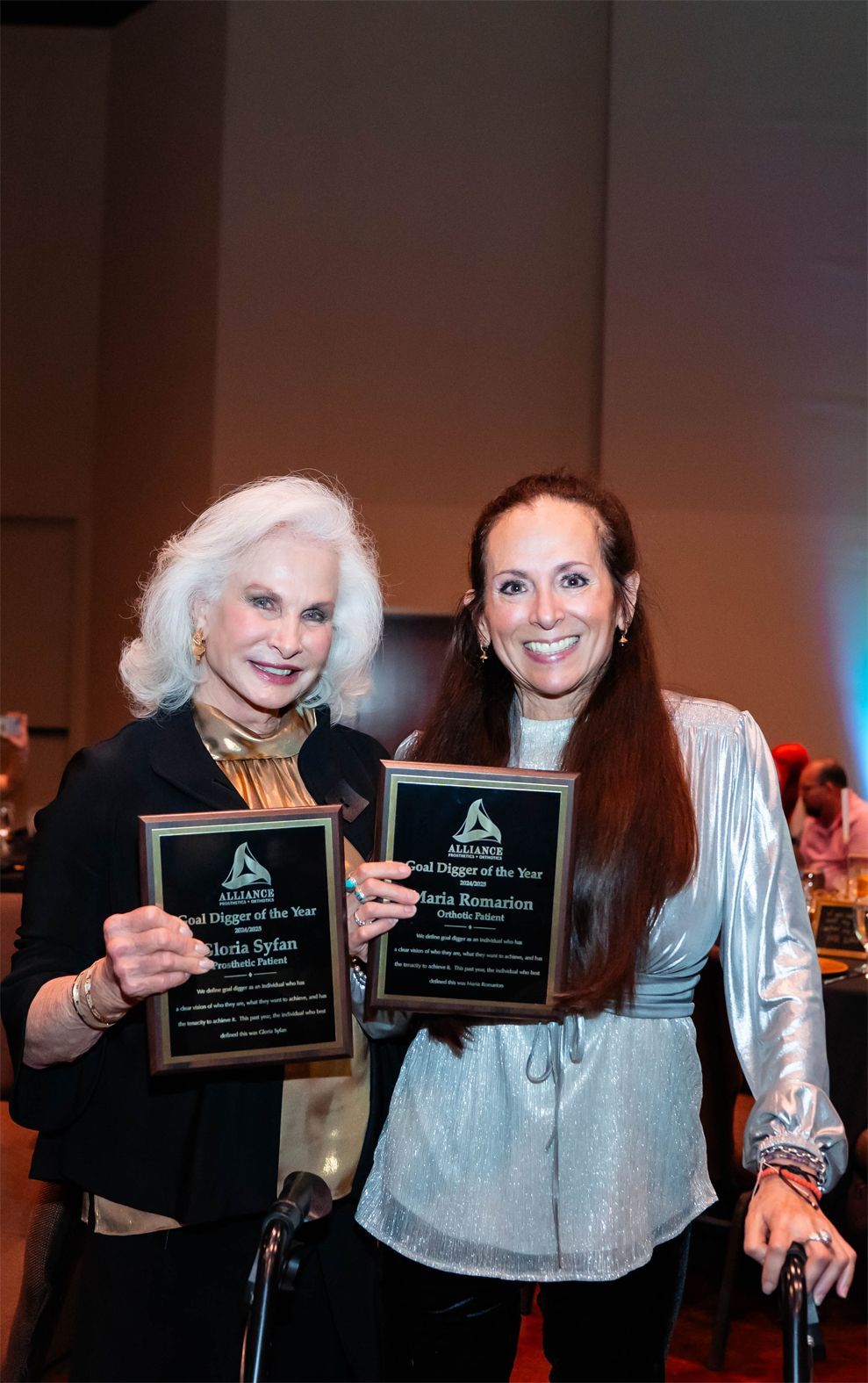Two women stand side-by-side smiling, holding matching award plaques at an indoor event.