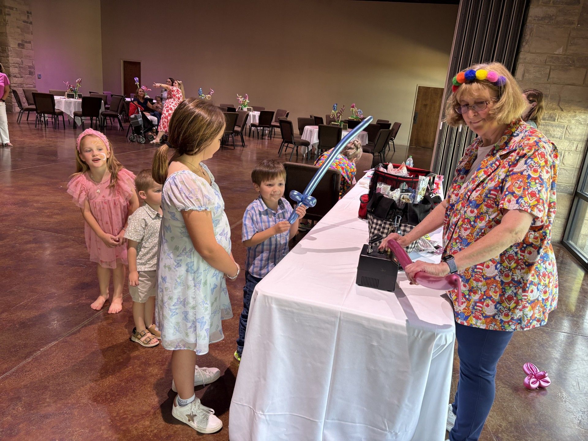 Children looking at a woman behind a table at an indoor event, with balloon swords and a table display.