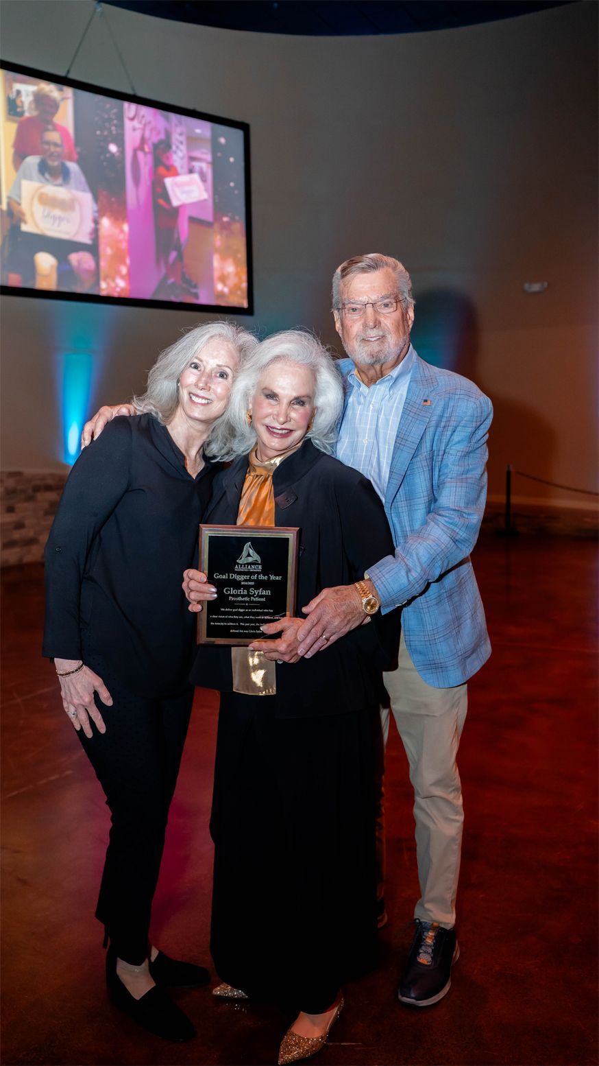 Three people stand closely together in a warmly lit room, smiling at the camera. The person in the center holds an award plaque. A projected image is visible in the background.