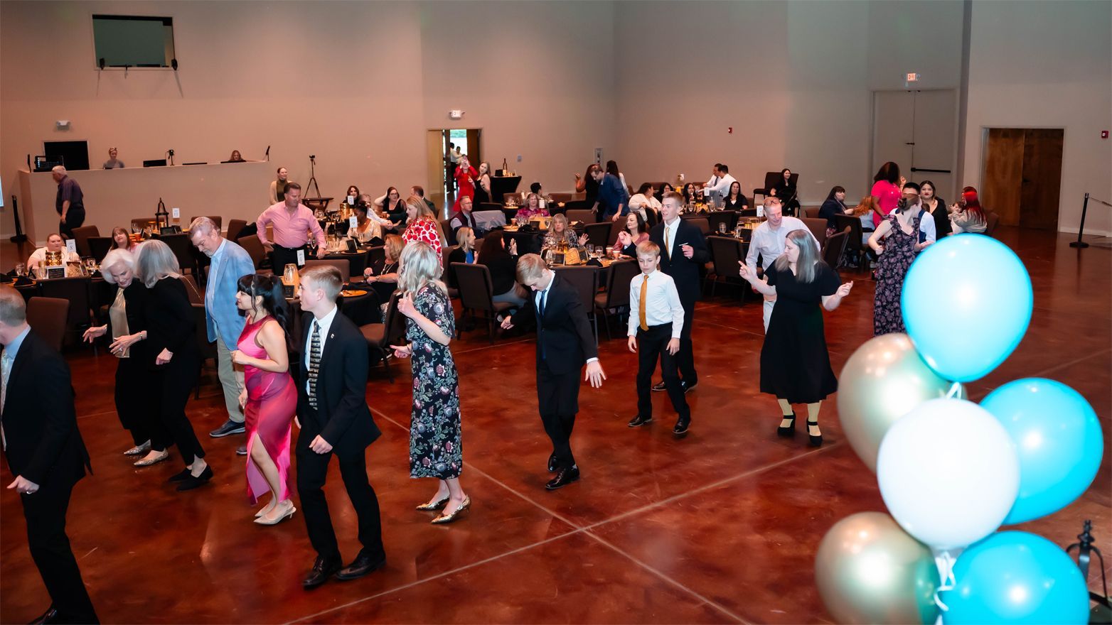 A group of people in formal attire dances together in a large event hall, with teal and white balloons in the foreground.