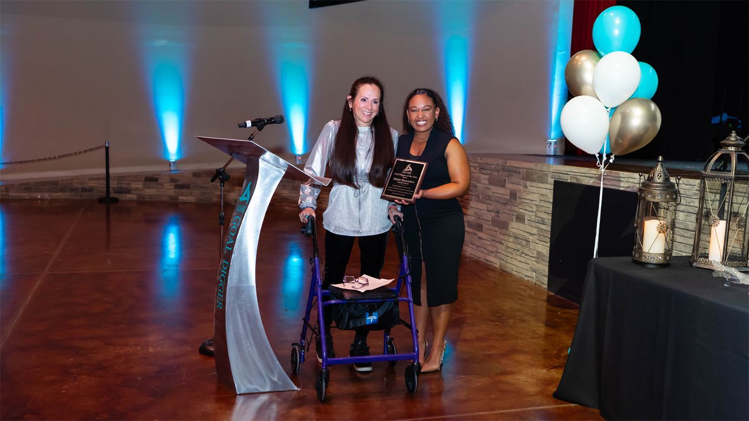 Two women smile on stage, one using a walker, holding an award plaque. The setting is softly lit with blue uplighting, balloons, and lanterns.
