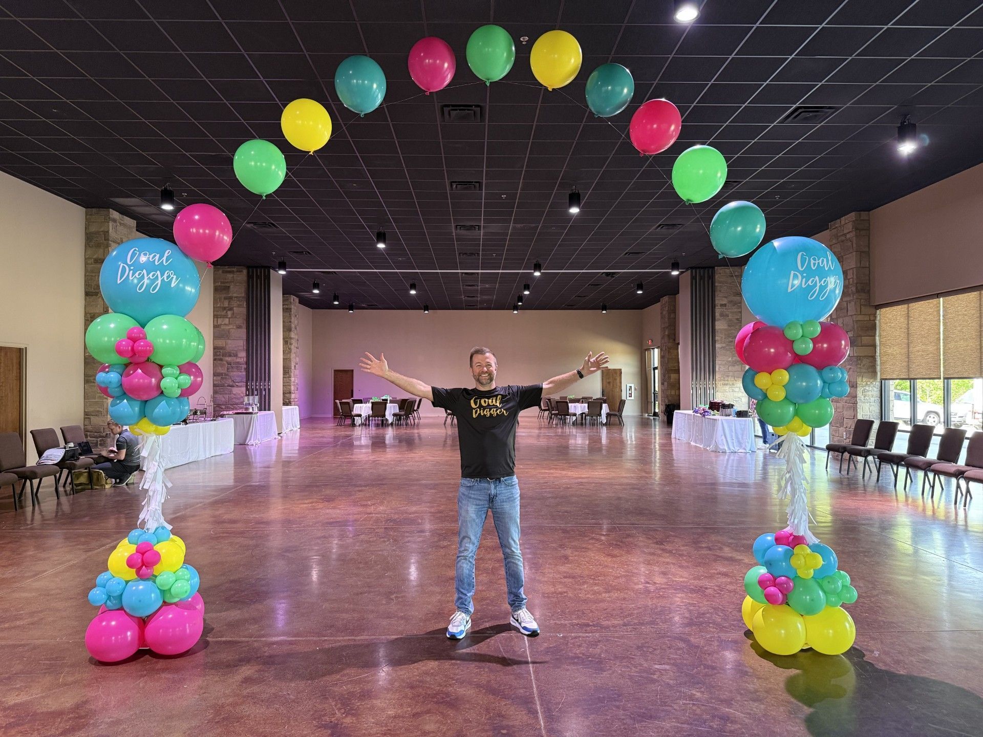 Person standing under a colorful balloon arch in a large empty hall with arms outstretched.