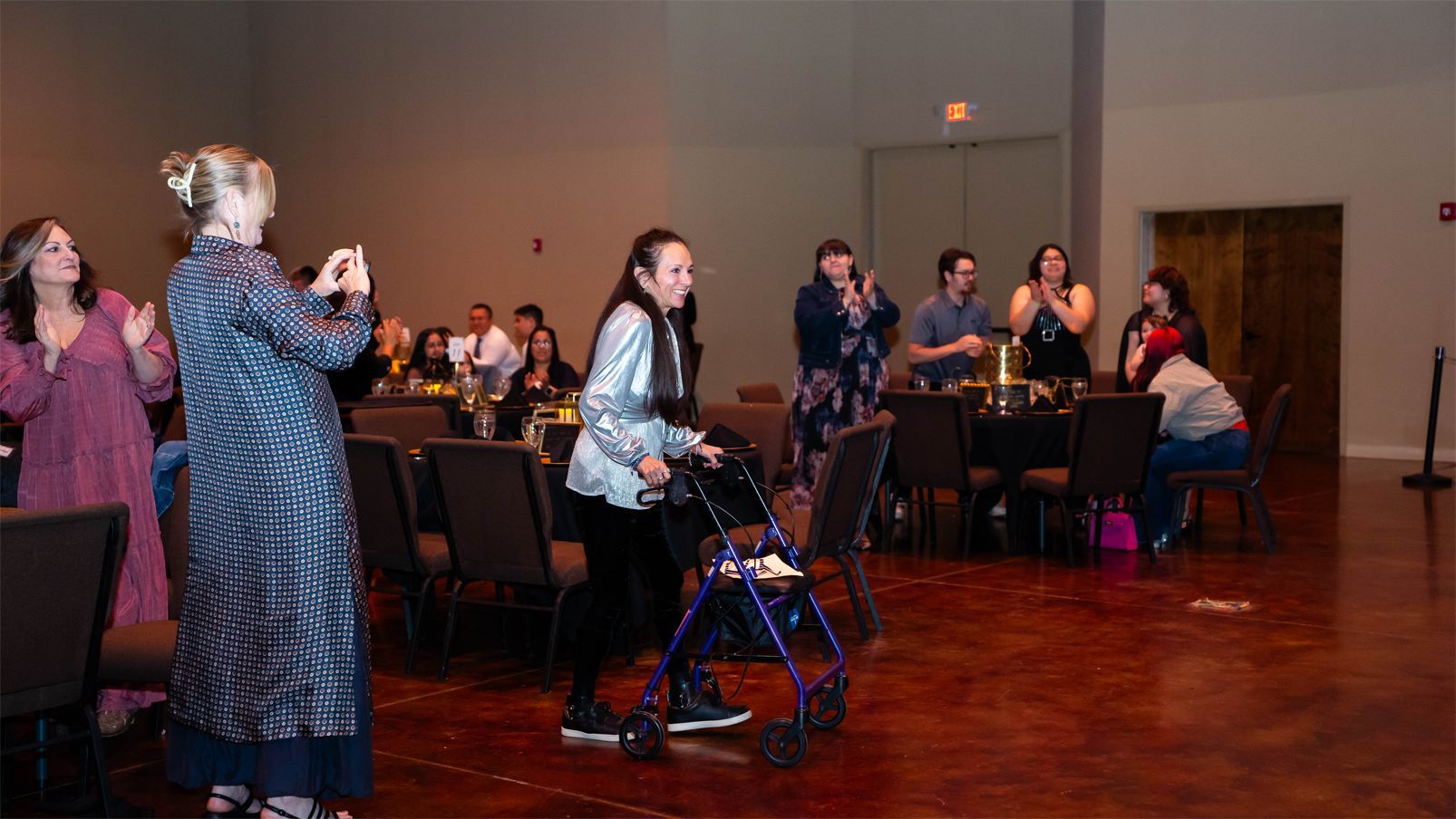 A woman with a walker moves across a dimly lit banquet hall, surrounded by clapping attendees in formal attire, creating a supportive atmosphere.