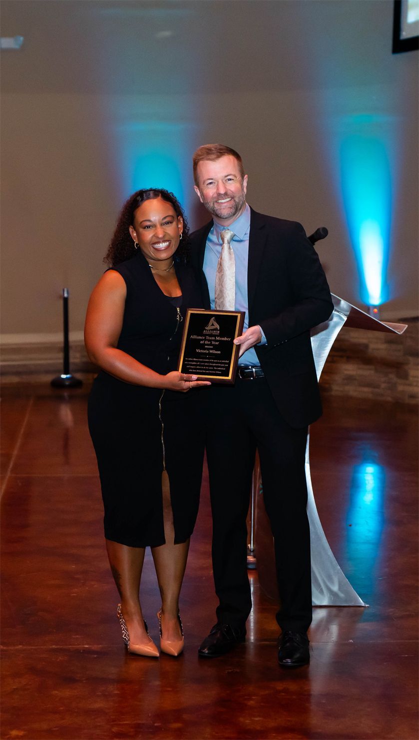 A person in a dark dress and a person in a suit smile while holding an award together on a stage with blue lighting.