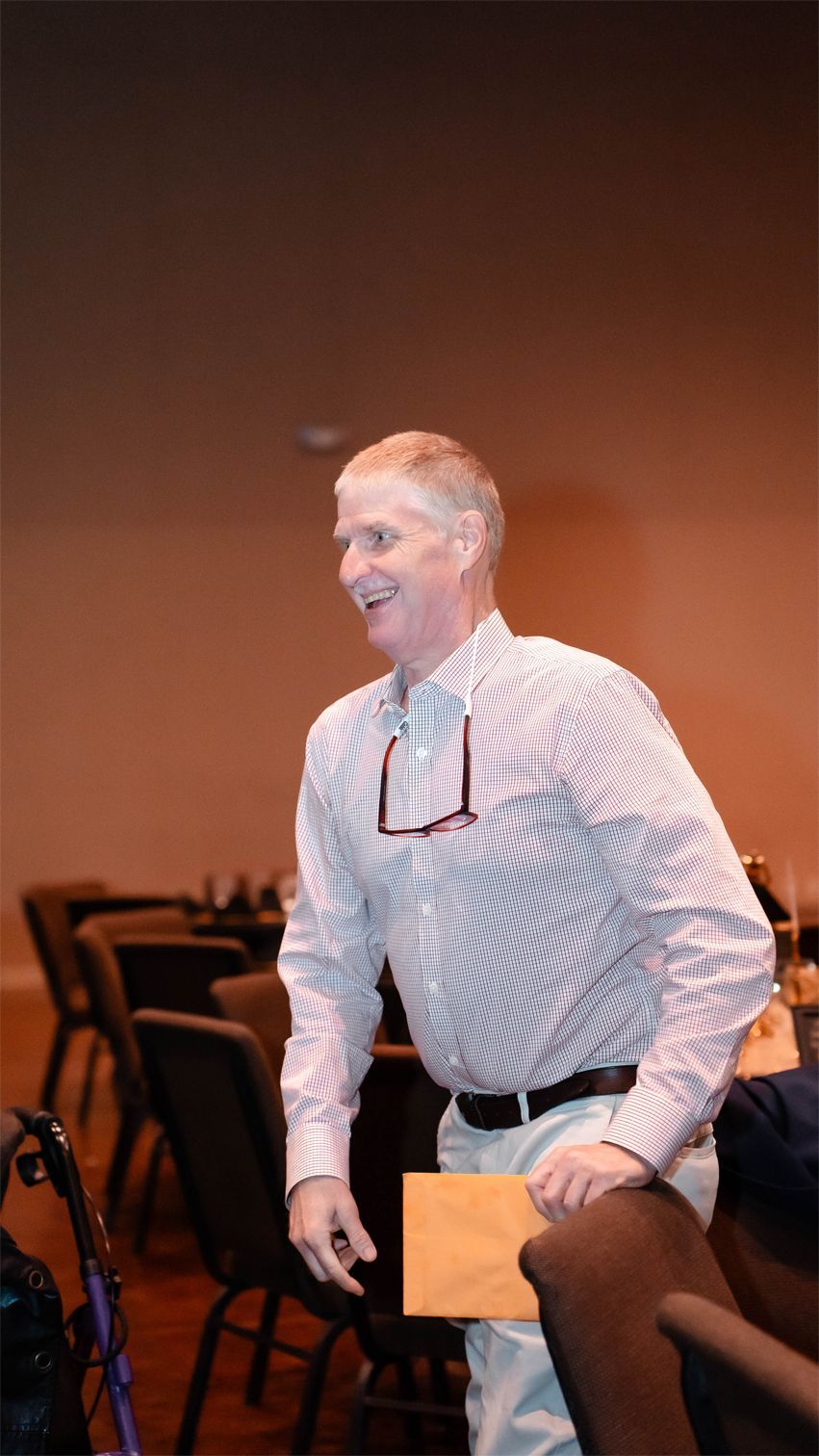 A smiling person in a button-down shirt and light-colored pants holds a tan envelope while standing in a room with chairs.