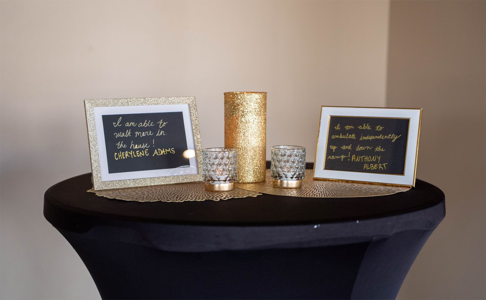 A black table displays two framed blackboards with gold borders and handwritten notes, two glass candle holders, and a tall gold glitter candle. Elegant and celebratory atmosphere.
