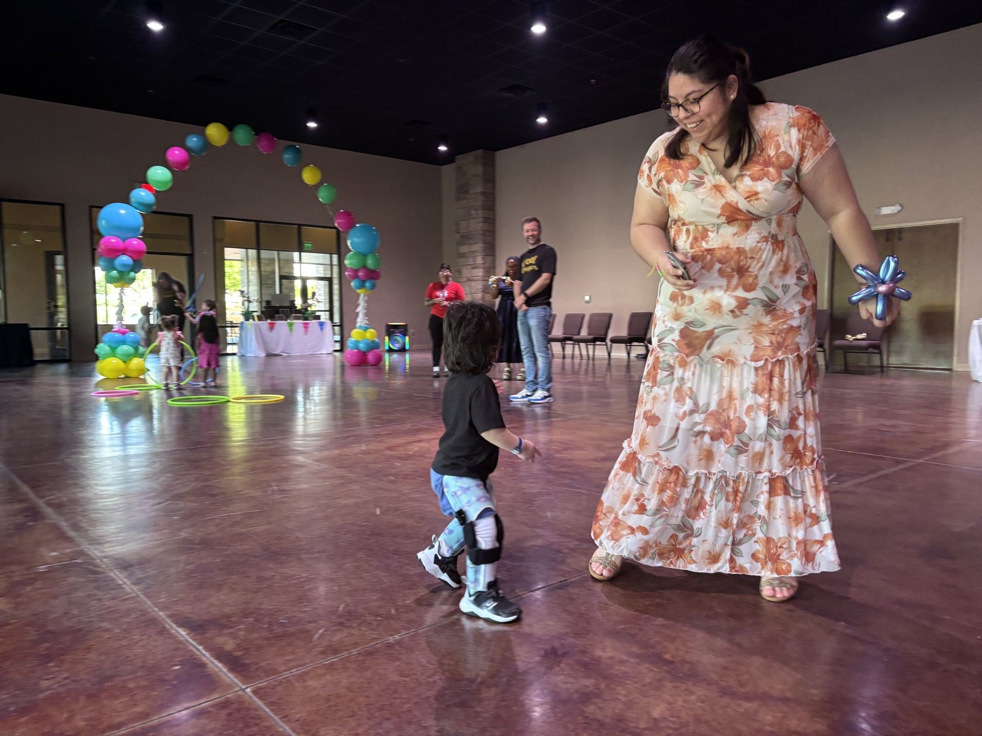 Child reaching toward woman in floral dress at an indoor event with colorful balloon arch