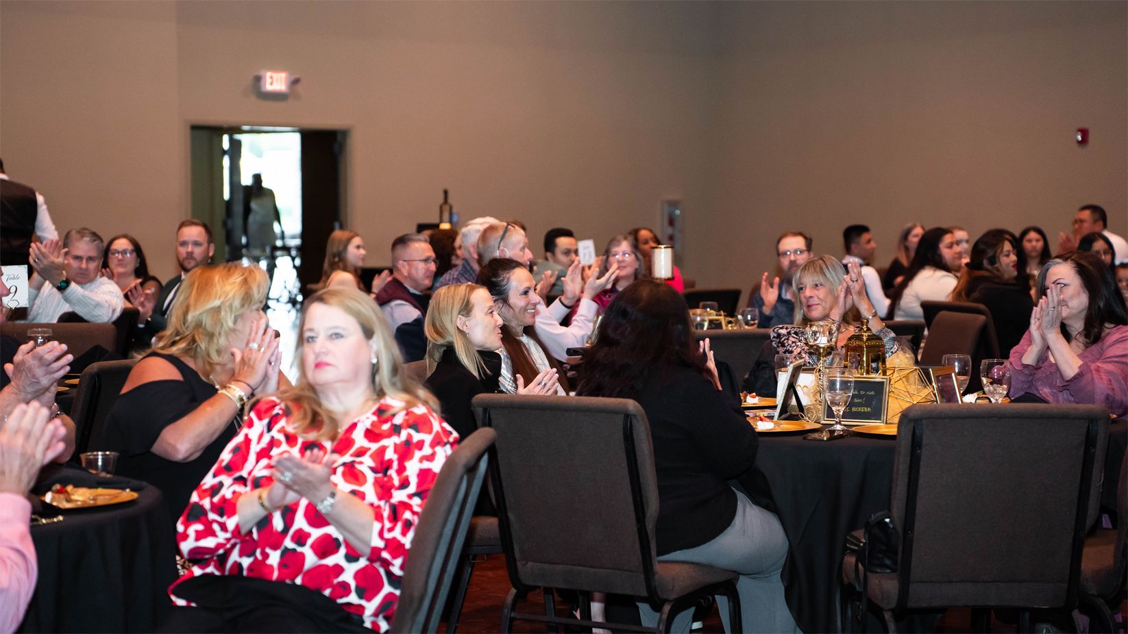A group of people seated at tables in a dimly lit conference room, clapping and smiling. The atmosphere is formal and celebratory.
