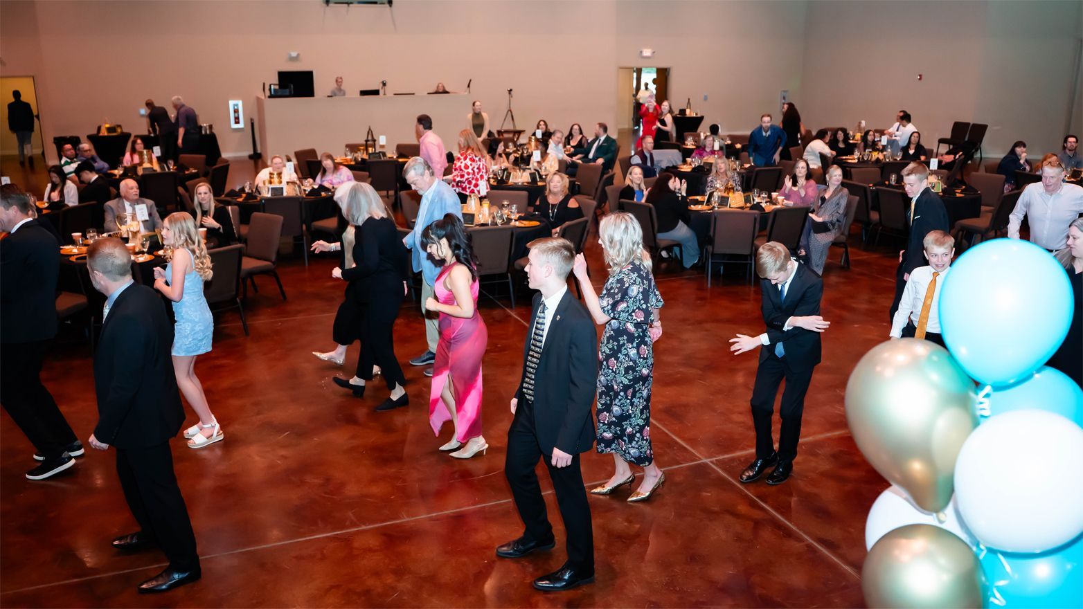 A ballroom dance floor filled with people in formal attire, with a cluster of blue and gold balloons in the foreground.