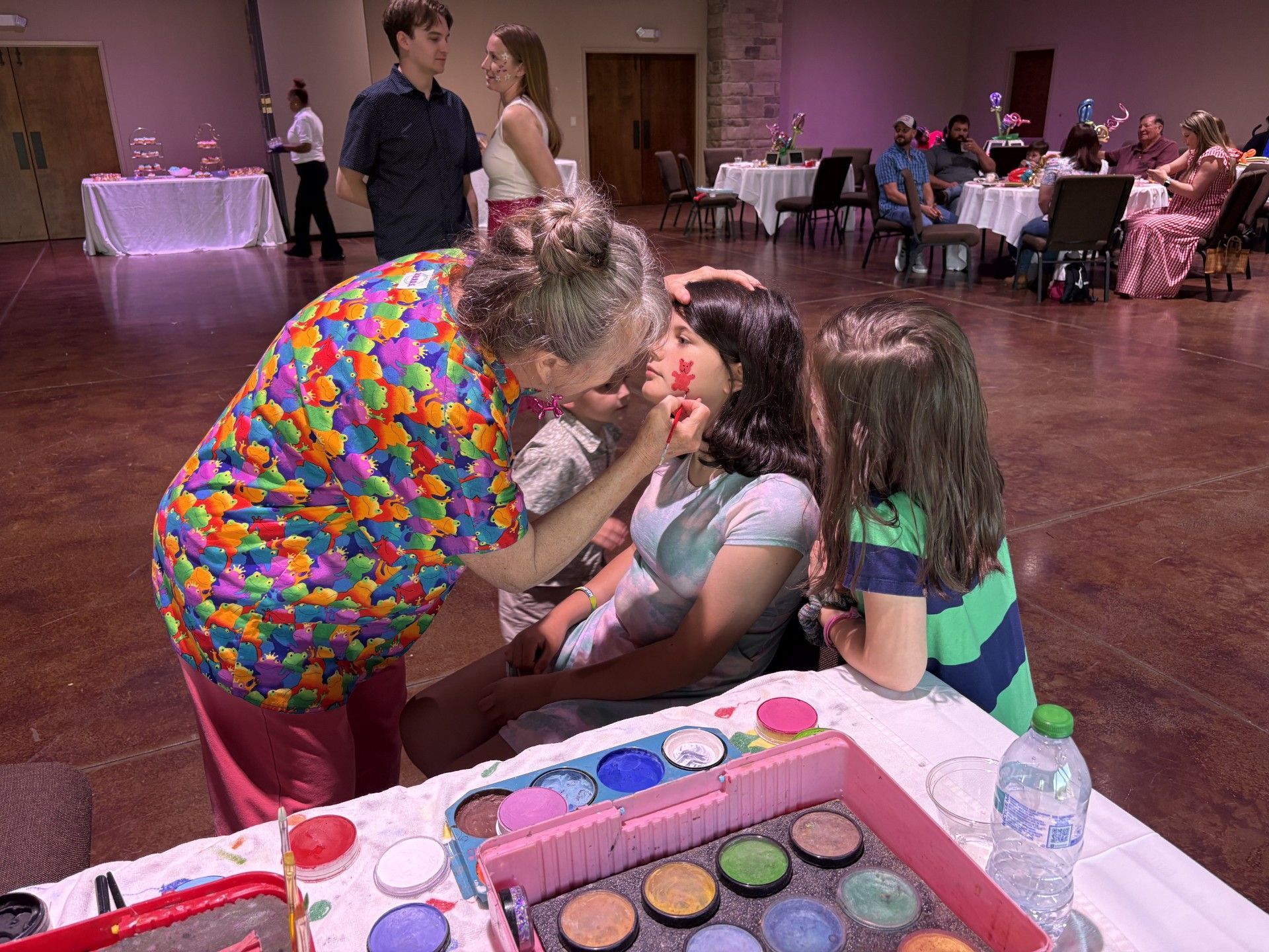 Woman applying face paint to a seated child at a party table with makeup supplies in the foreground.