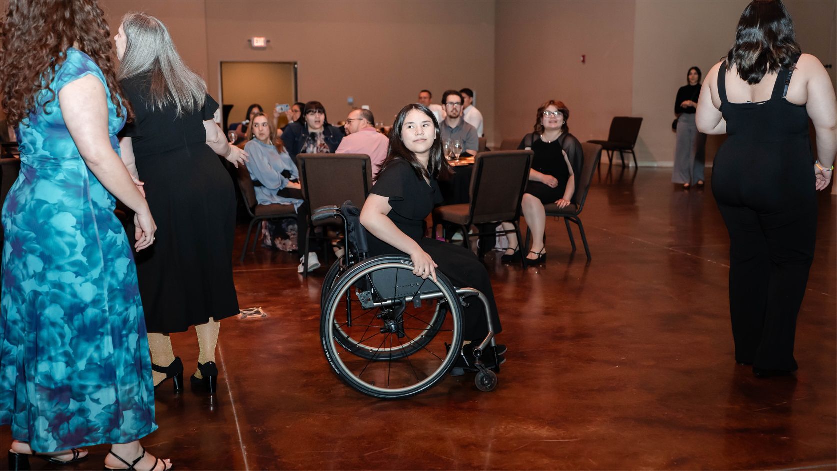 People in formal wear gather in a large room; one person in a wheelchair sits in the center while others stand or sit.