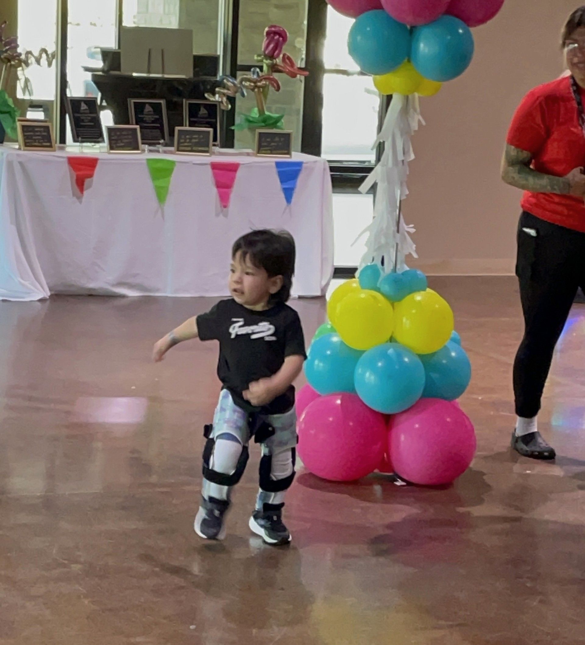 Toddler in leg braces stands in a decorated indoor hall beside a colorful balloon column.