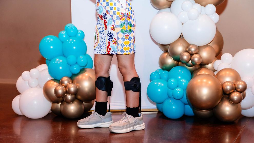 A person wearing leg braces and sneakers stands between two decorative balloon arches featuring teal, gold, and white.