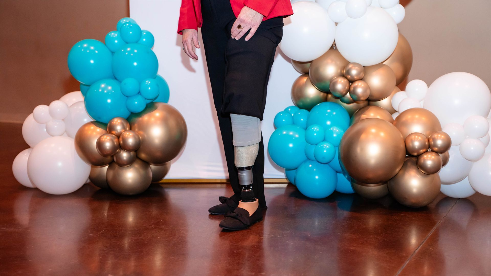 A person wearing a prosthetic lower leg stands between clusters of teal, gold, and white balloons.