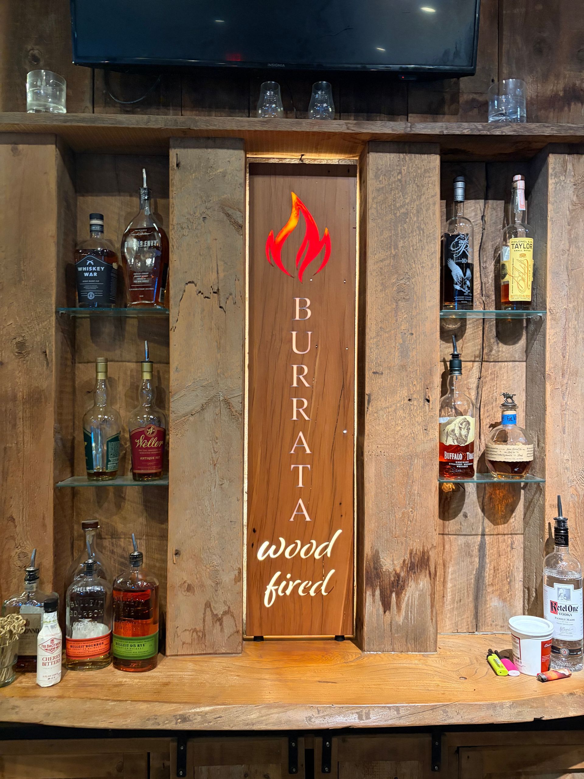 Wood-fired grill with rustic wooden shelves and bottles, featuring a red flame logo and 