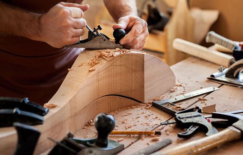 Hands carving wood with a hand plane on a workbench, surrounded by wood shavings and tools