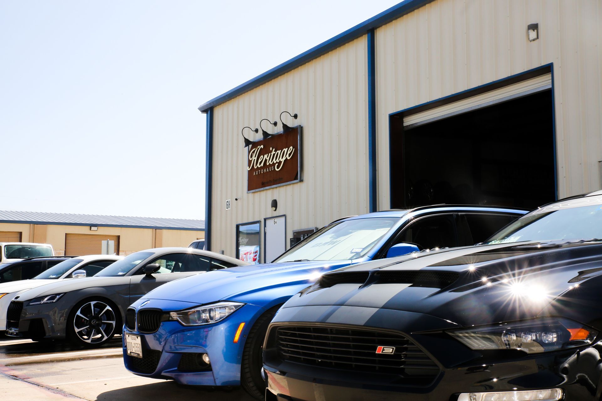 Several parked sports cars outside a garage, including a black car in the foreground and a blue car behind it