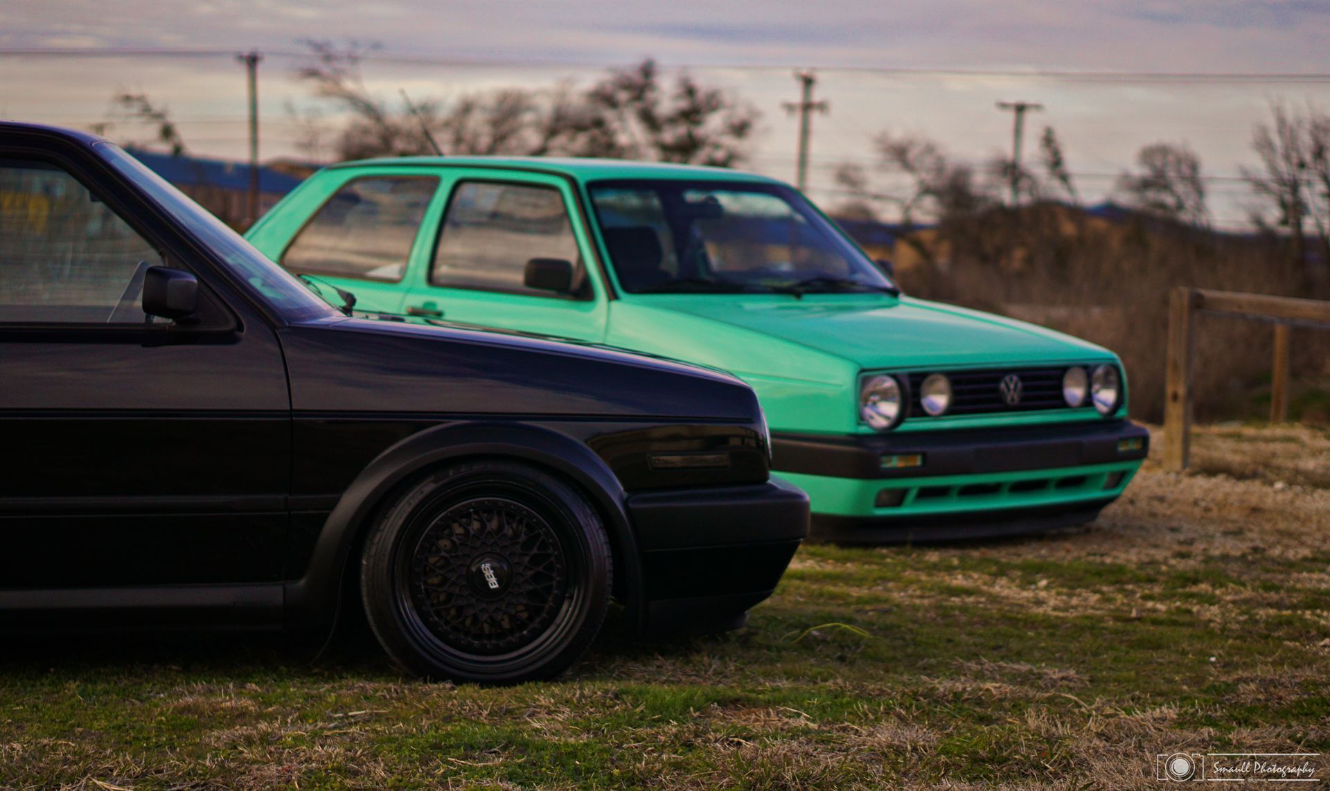 Green hatchback beside a black car in a grassy field under an overcast sky