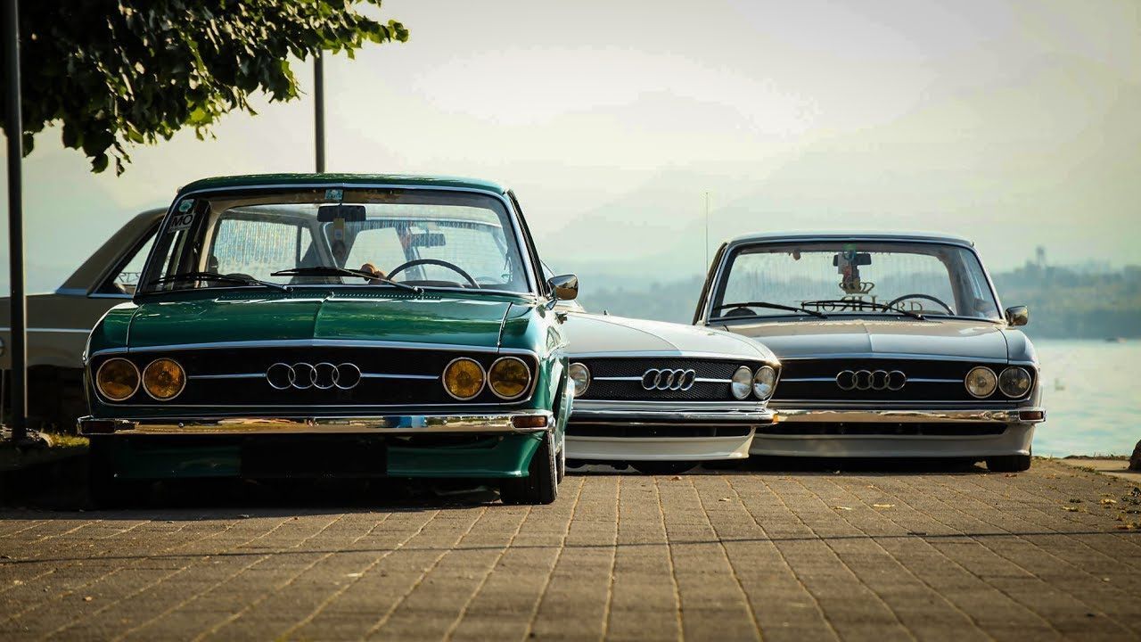 Two classic cars parked side by side on a paved road at sunset, one green and one dark gray