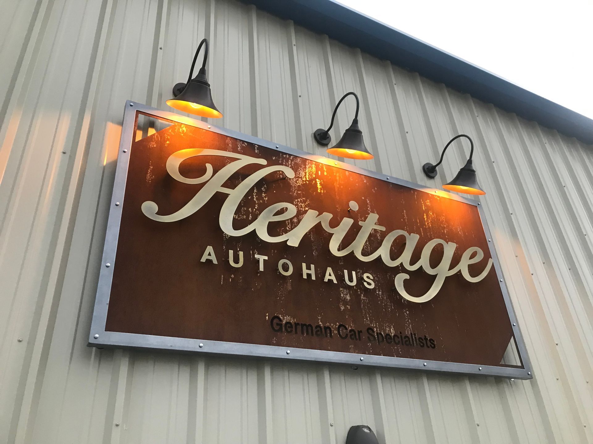 Heritage Autohaus sign on a corrugated metal building, lit by three lamps at dusk