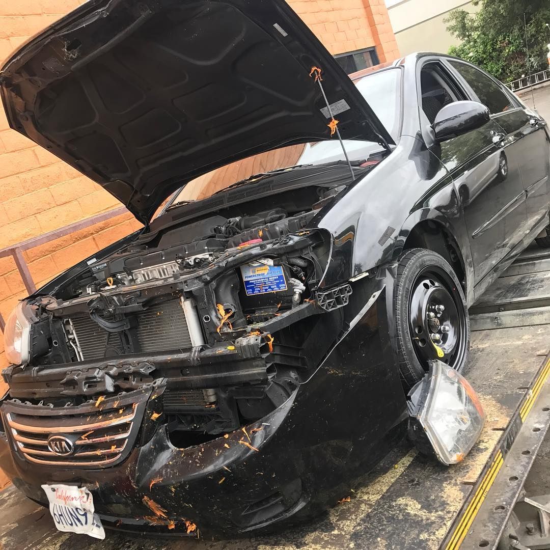 Black car with front-end damage, hood open, headlight detached. On a tow truck, exterior setting.