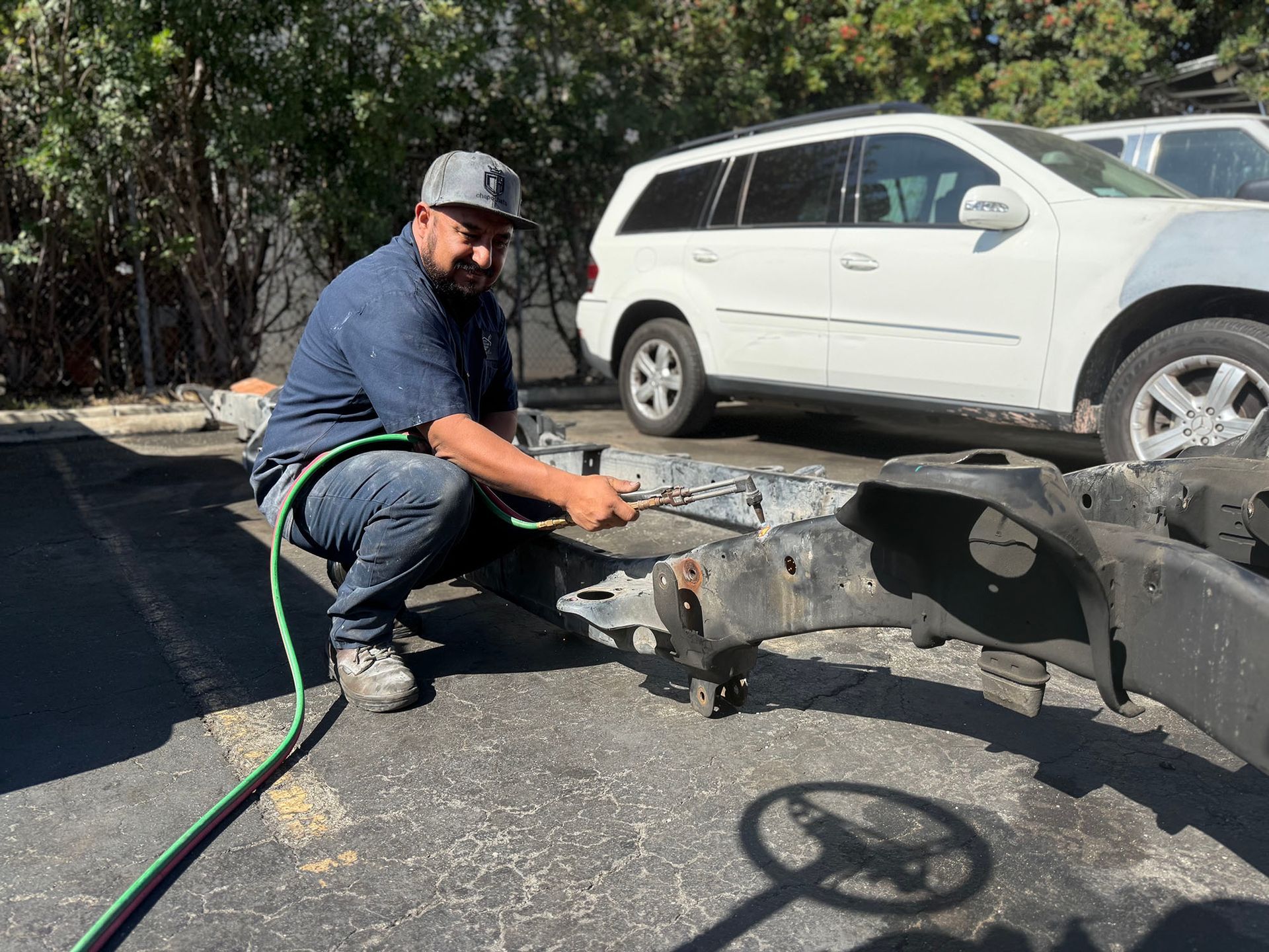 Man in work clothes cleans a car frame with a hose outdoors near a white SUV.