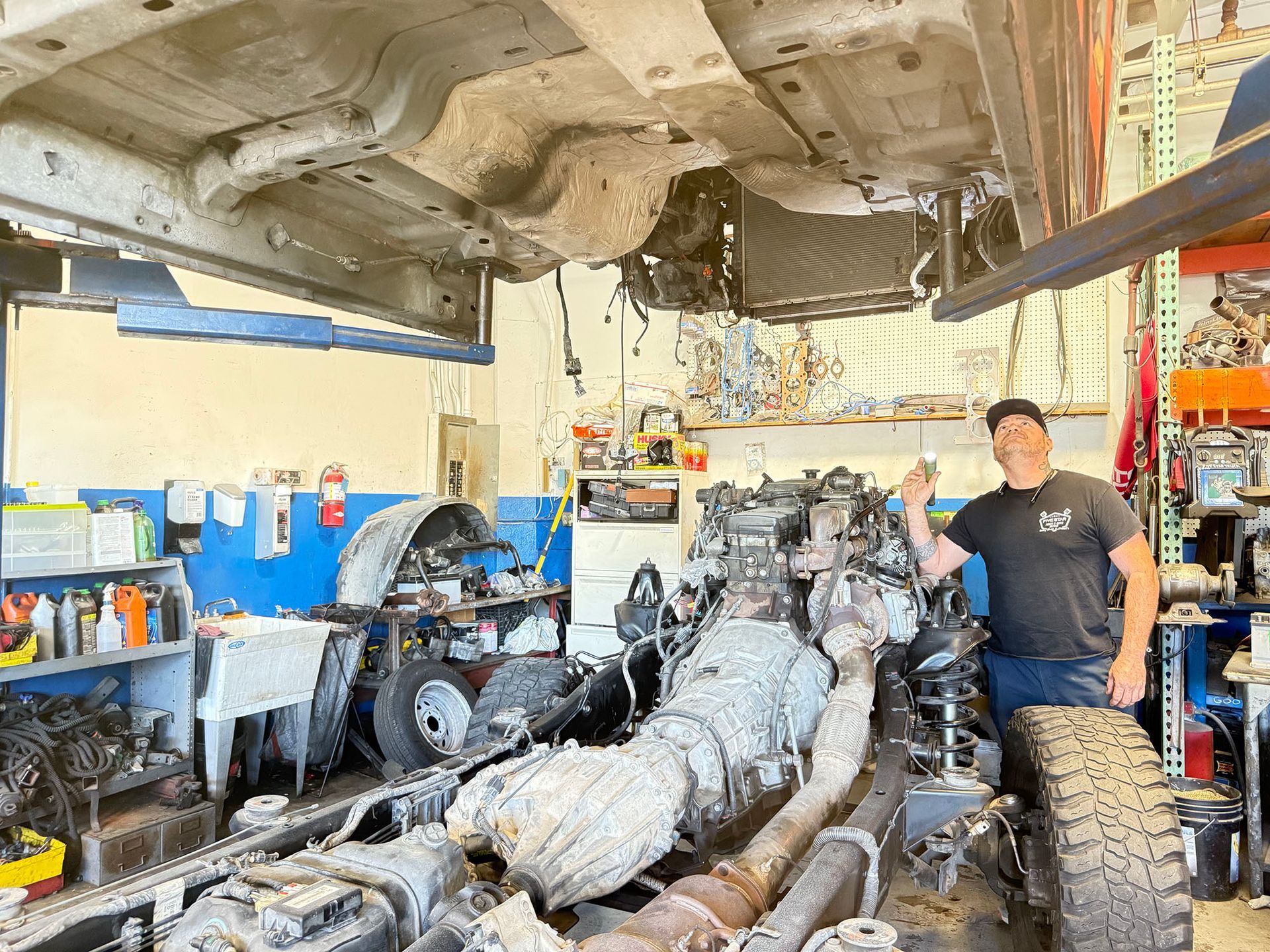 Mechanic in a shop pointing towards a truck chassis. Underneath vehicle. Workshop setting with parts.