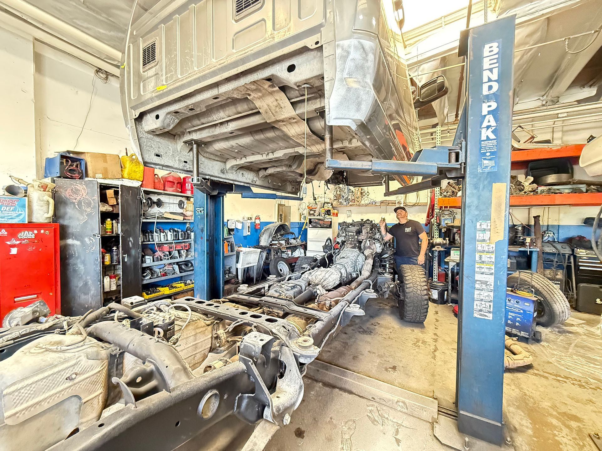 A truck chassis is being worked on in a garage. A mechanic stands near the vehicle lift.