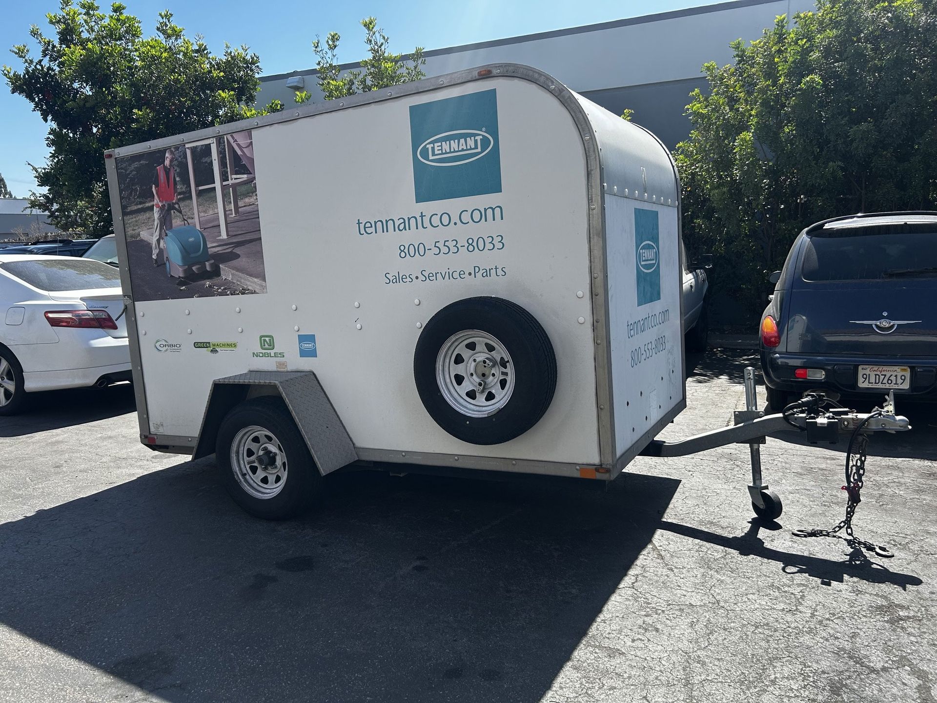 White utility trailer with logo and spare tire, parked on asphalt.