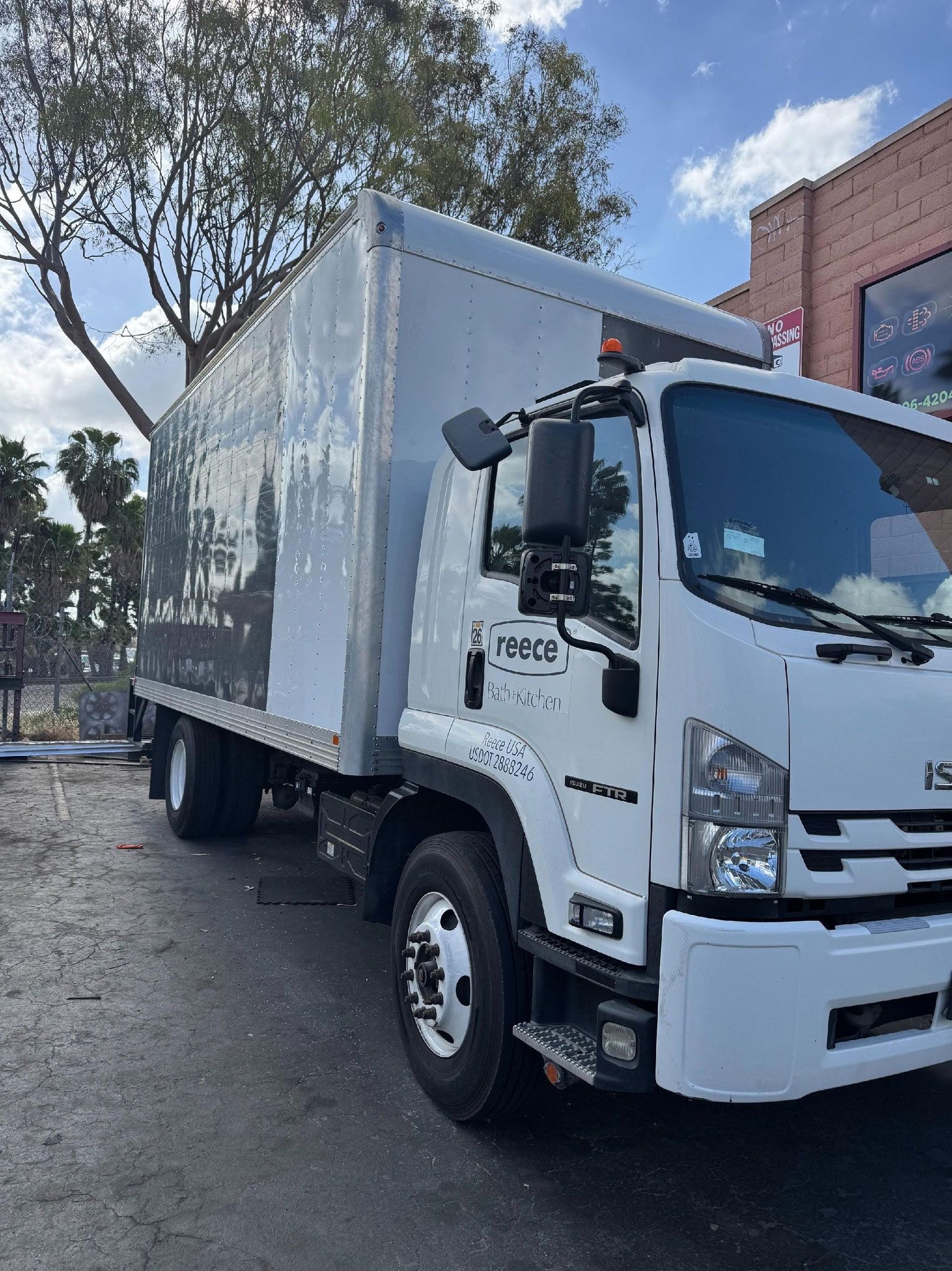 White Isuzu box truck parked on a street in front of a building with trees overhead.