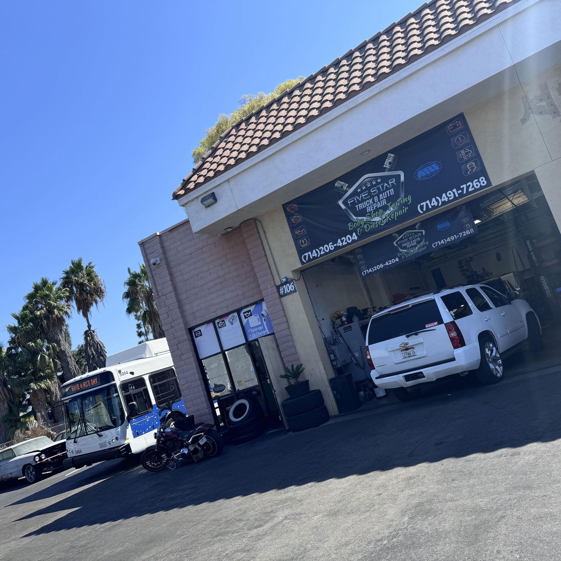A car service shop with a white SUV parked. A bus and a motorcycle are visible nearby.