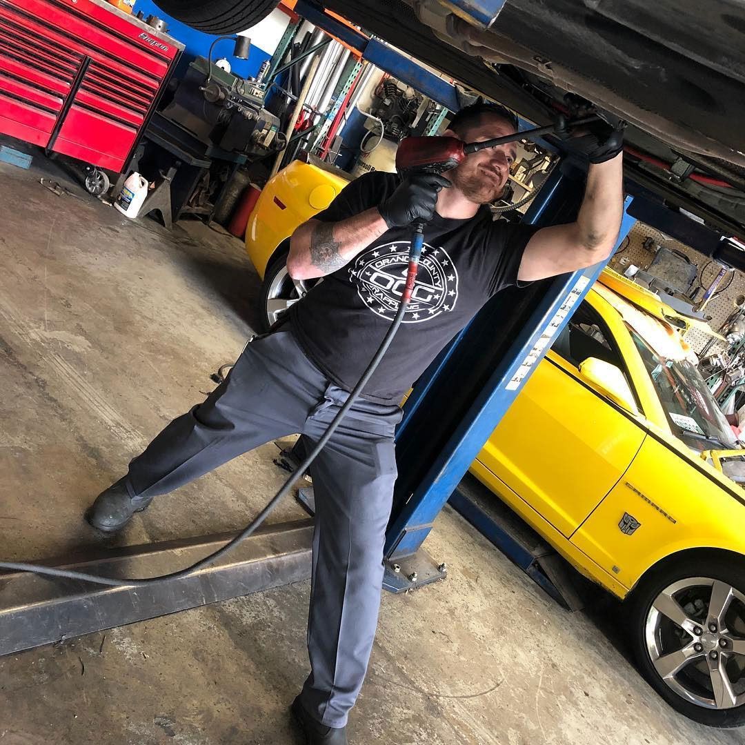 Mechanic working on the underside of a yellow car, using a tool. He's wearing a black shirt, gloves, and ear protection.