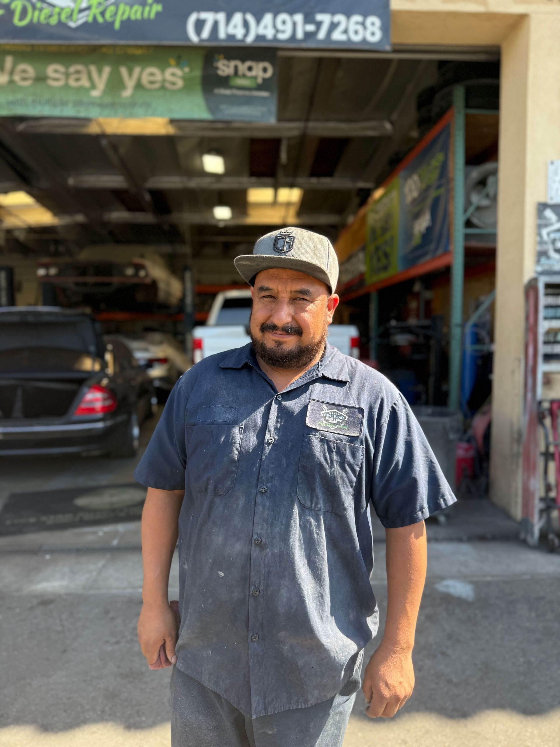 Mechanic stands at auto shop entrance. He's wearing a blue work shirt and a baseball cap. Shop sign has phone number.
