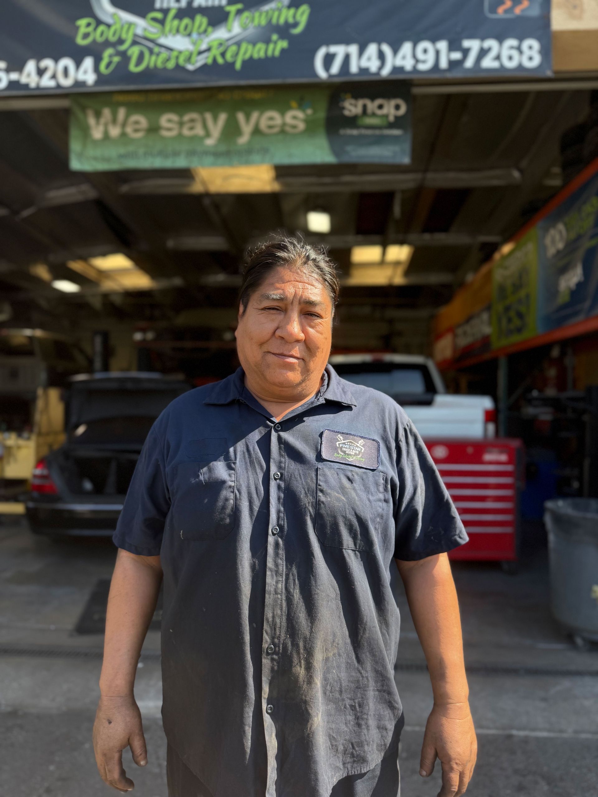 Man in blue work shirt stands in front of a body shop with a sign 