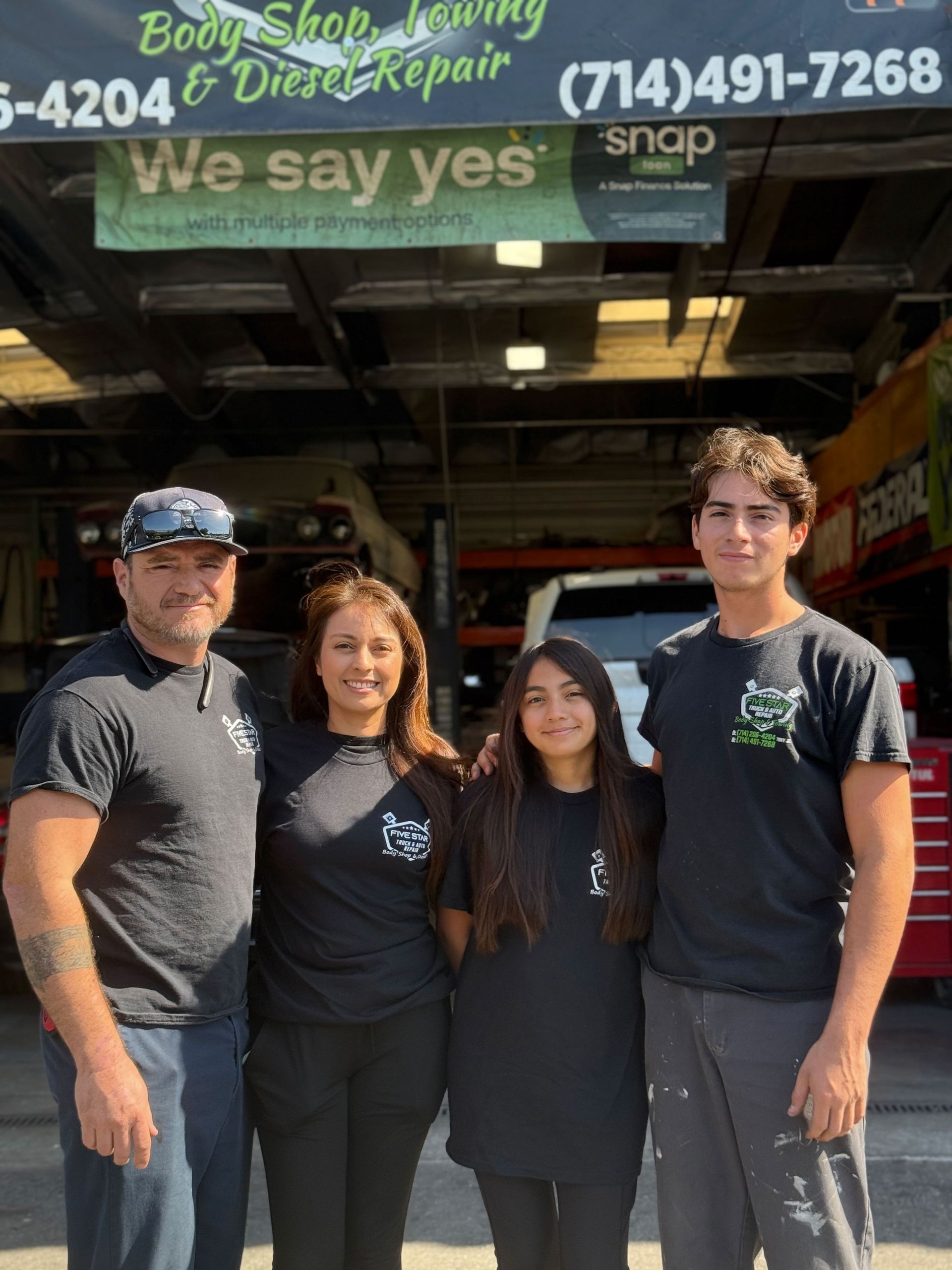 Four people in black shirts pose in front of a body shop, under a sign that reads 