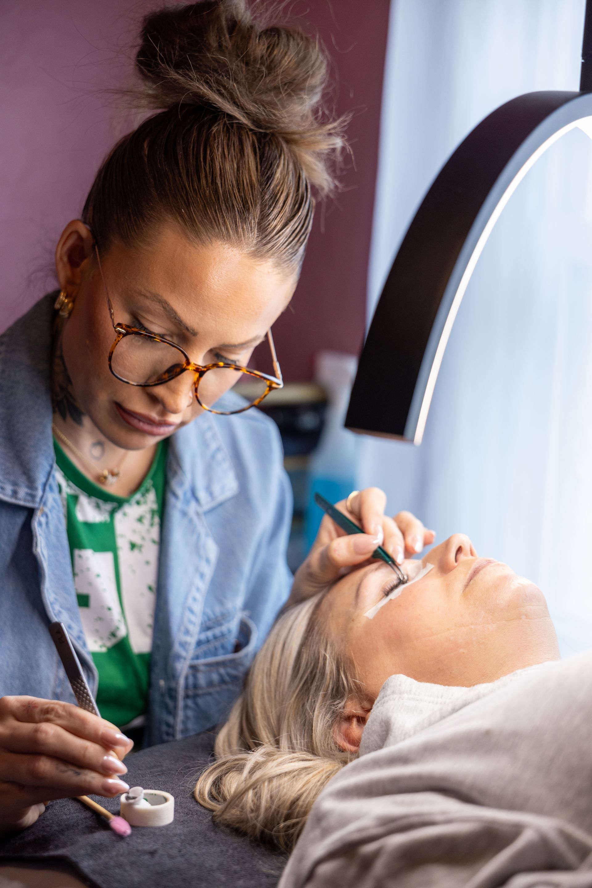 Eyebrow technician shaping a client’s brows in a salon chair near a window