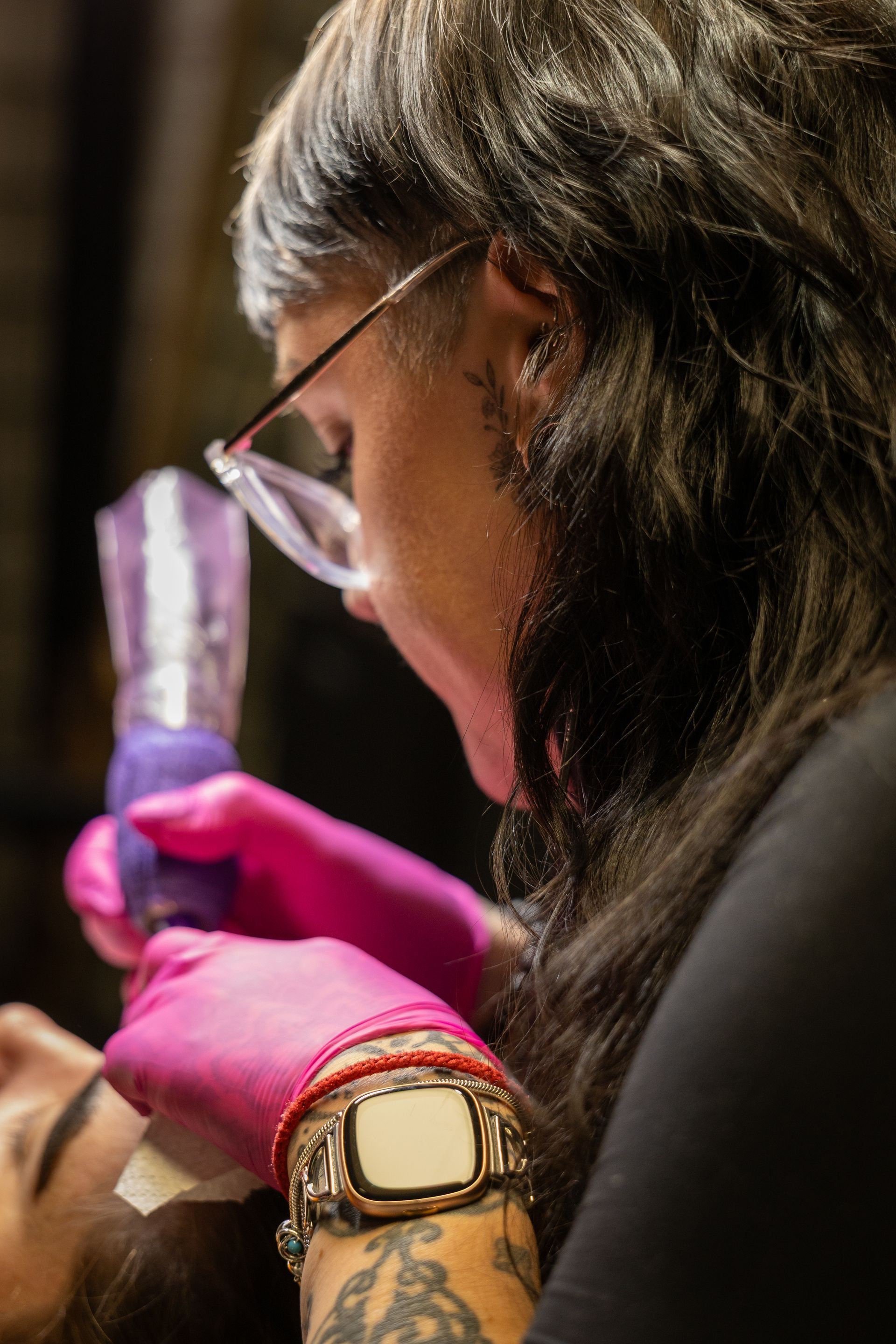 Gloved person examining a specimen under a microscope, wearing glasses and a gold watch