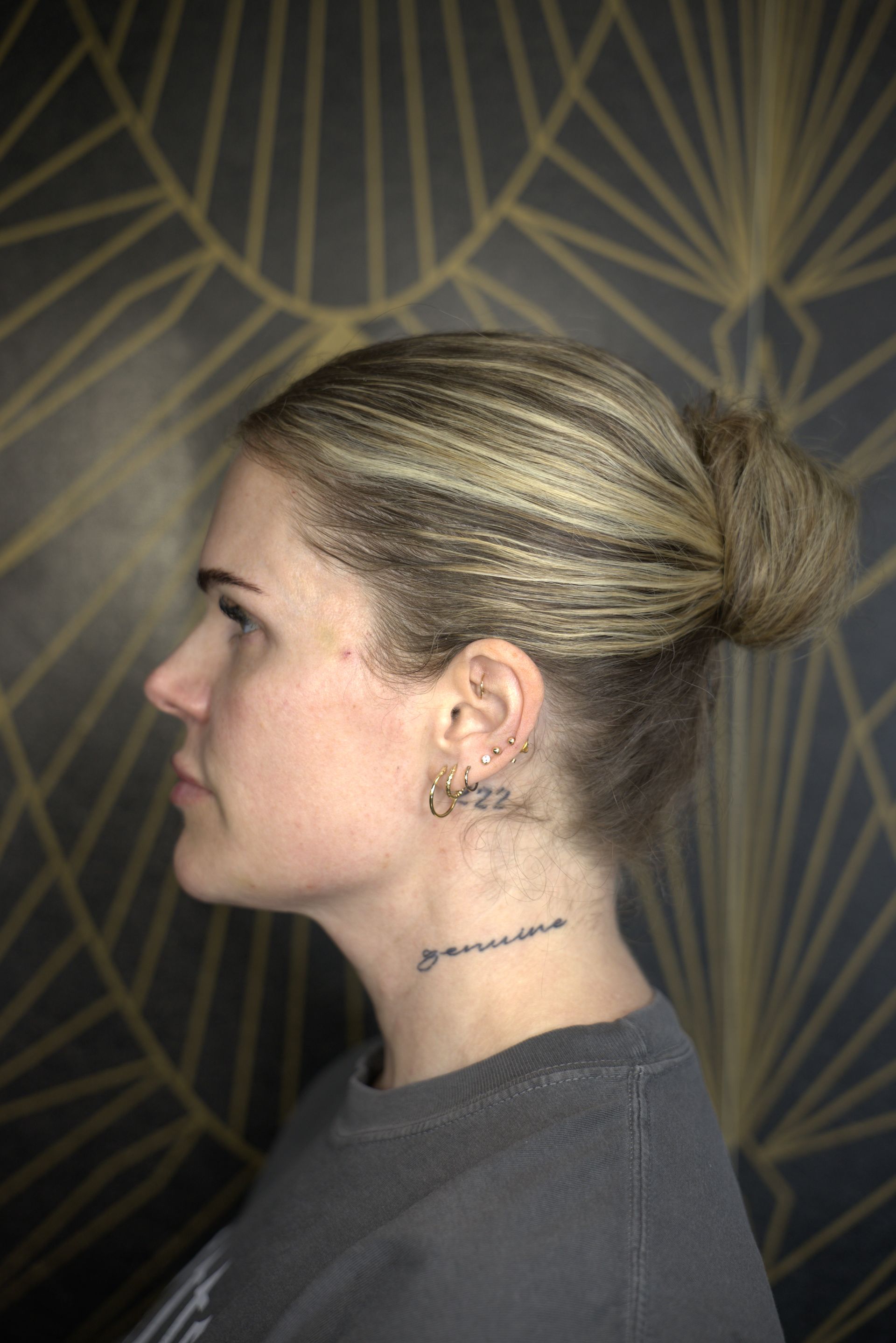 Profile view of a blonde woman with hair in a bun, wearing a gray top against a geometric wallpaper background