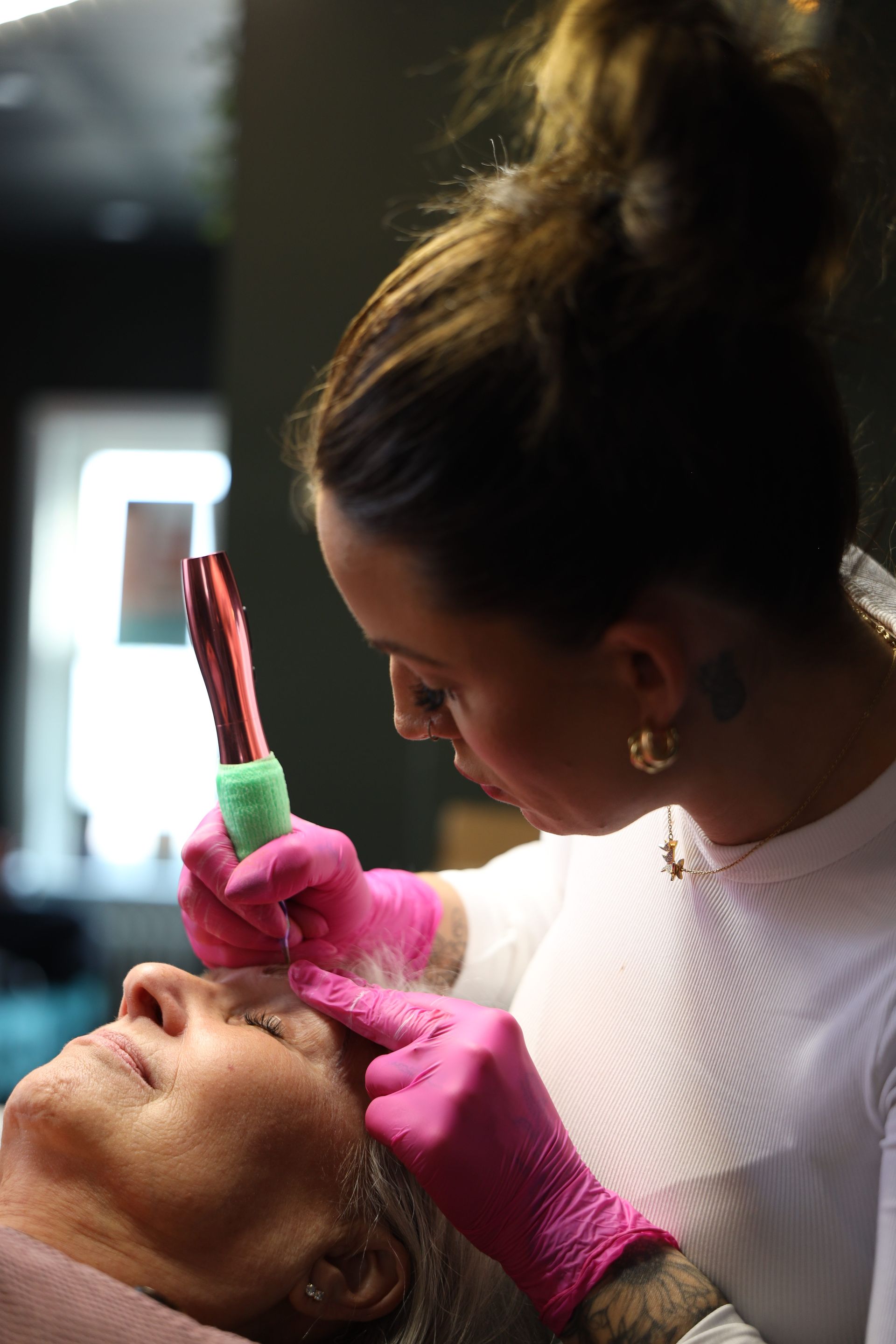 A cosmetologist applies makeup to a client lying down, using a brush near the client's eye.