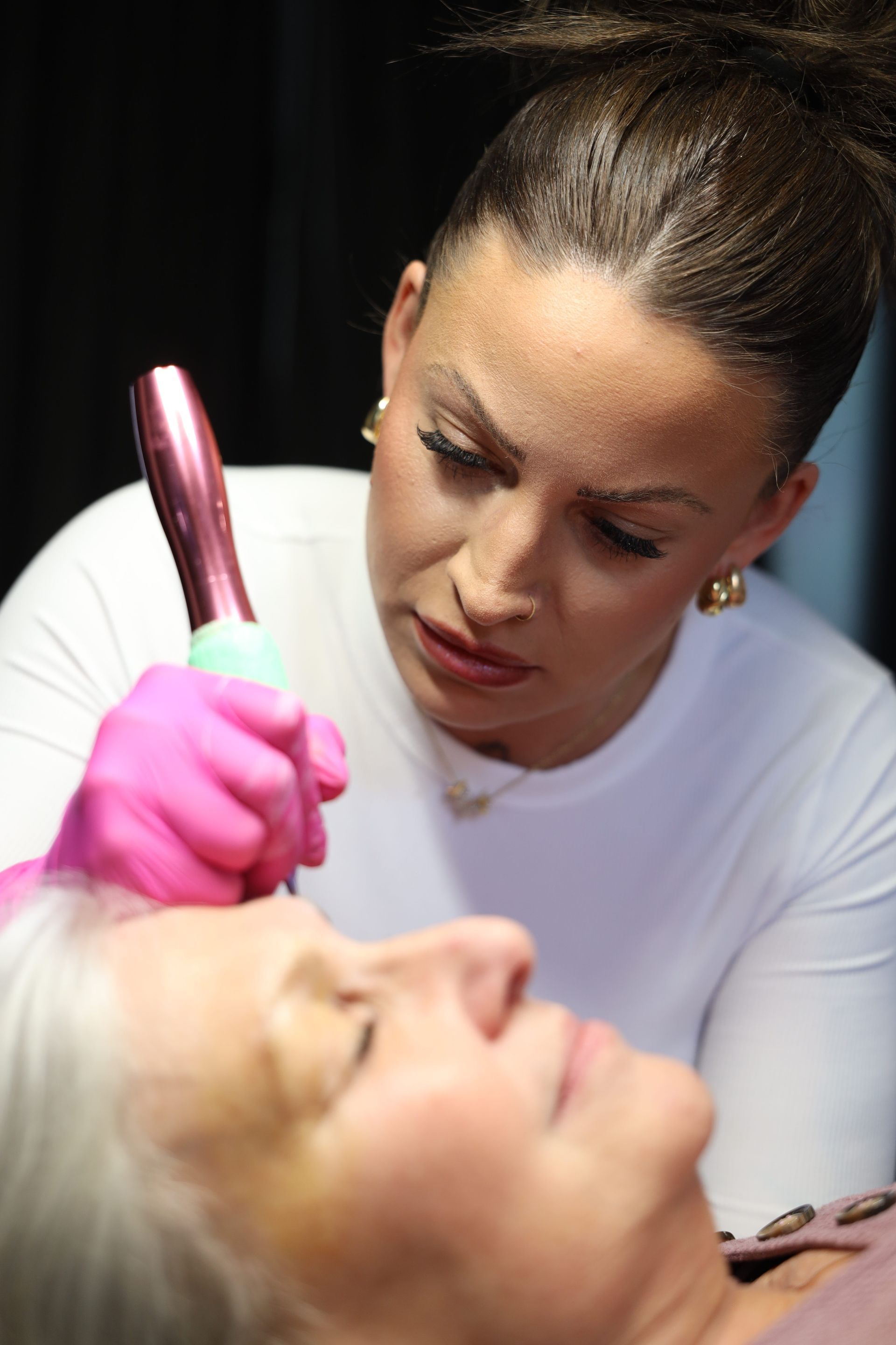A makeup artist applies makeup to a client’s face with a brush, wearing pink gloves.