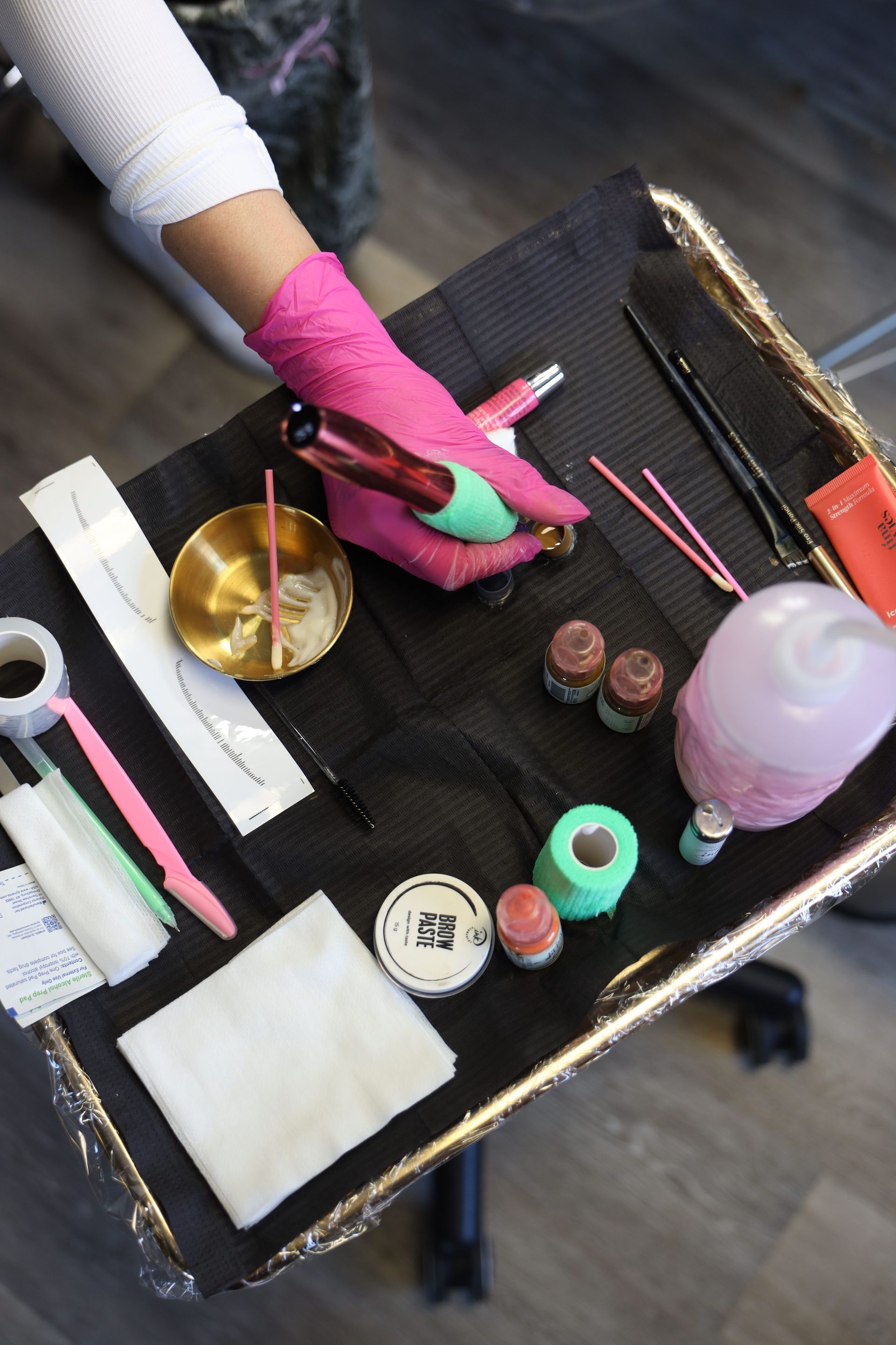 Gloved hand arranging manicure tools on a black tray, with nail polish, brushes, and a bowl.