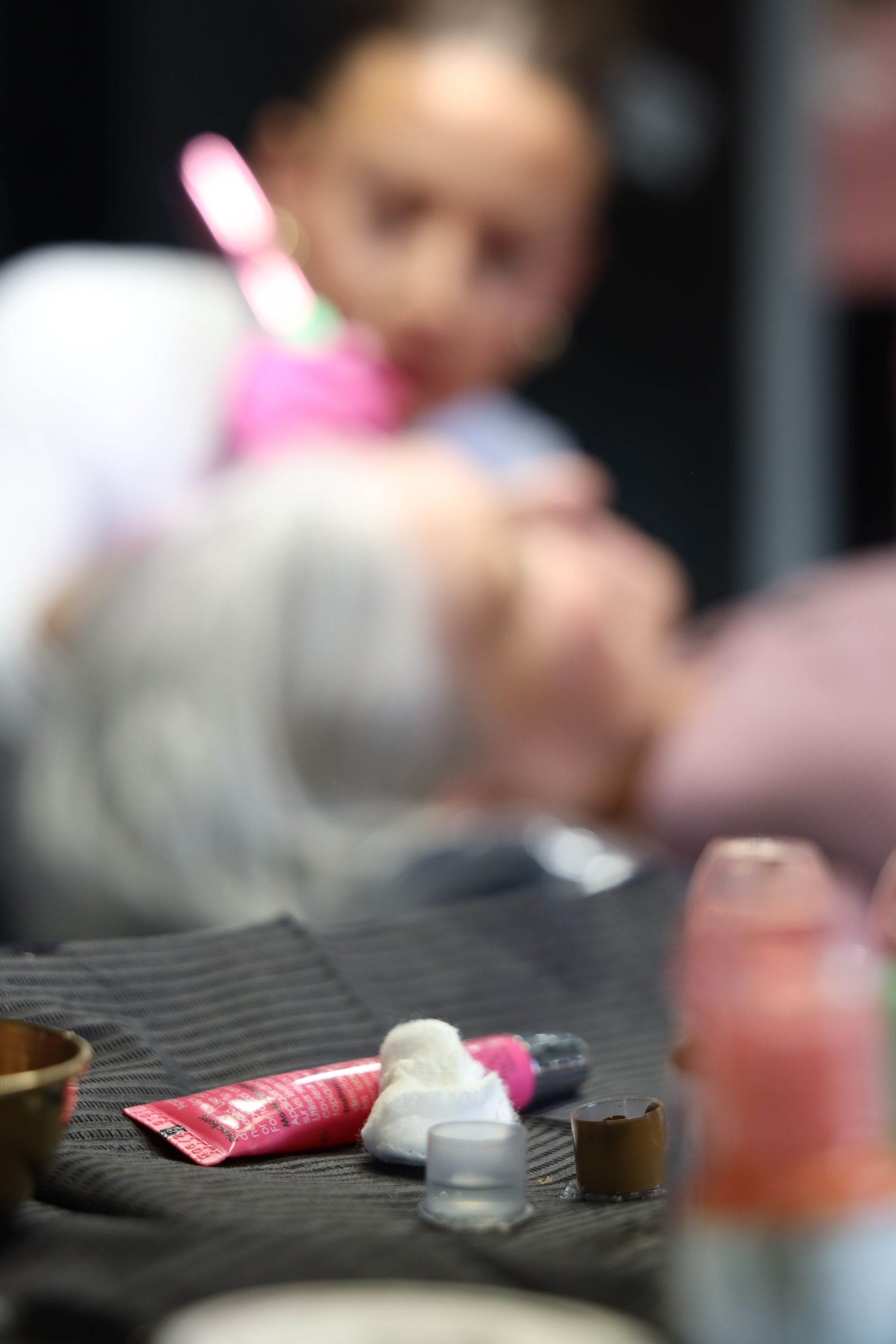 Blurred close-up of a tabletop with small cosmetic items and a pink tube in the foreground.