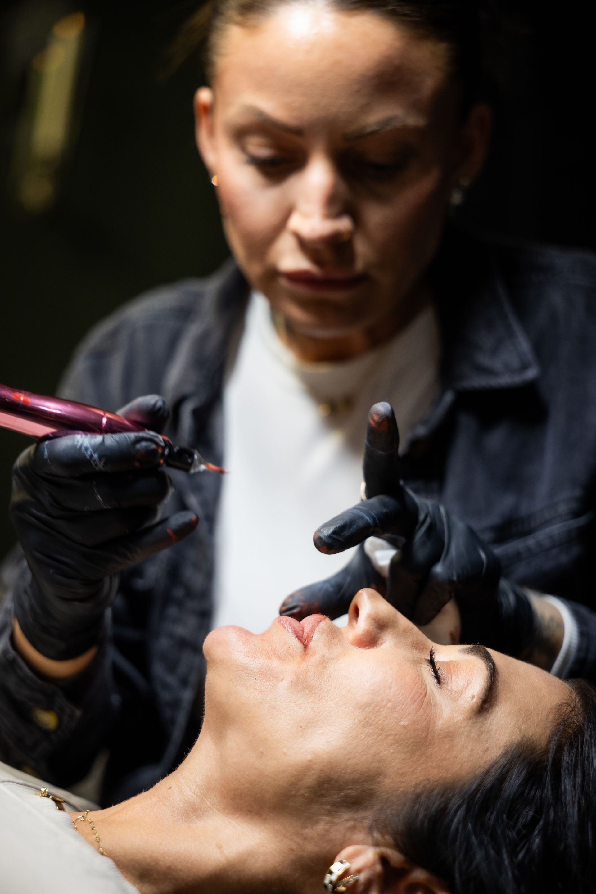 Technician applying lip treatment to a client lying back, with gloved hands and tools in a dim studio