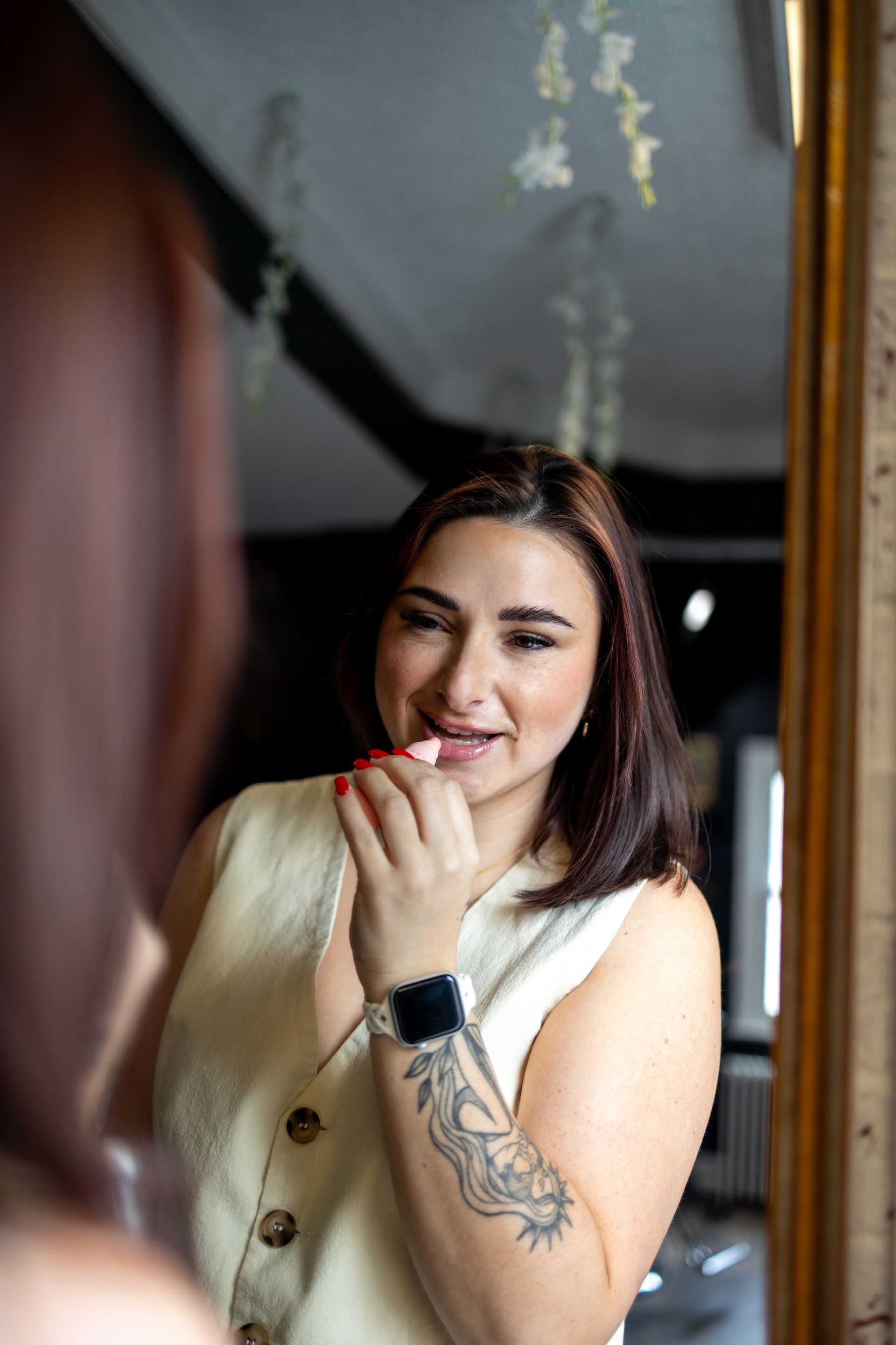 Woman smiling at a mirror, touching her lip with a hand, wearing a white top and smartwatch.