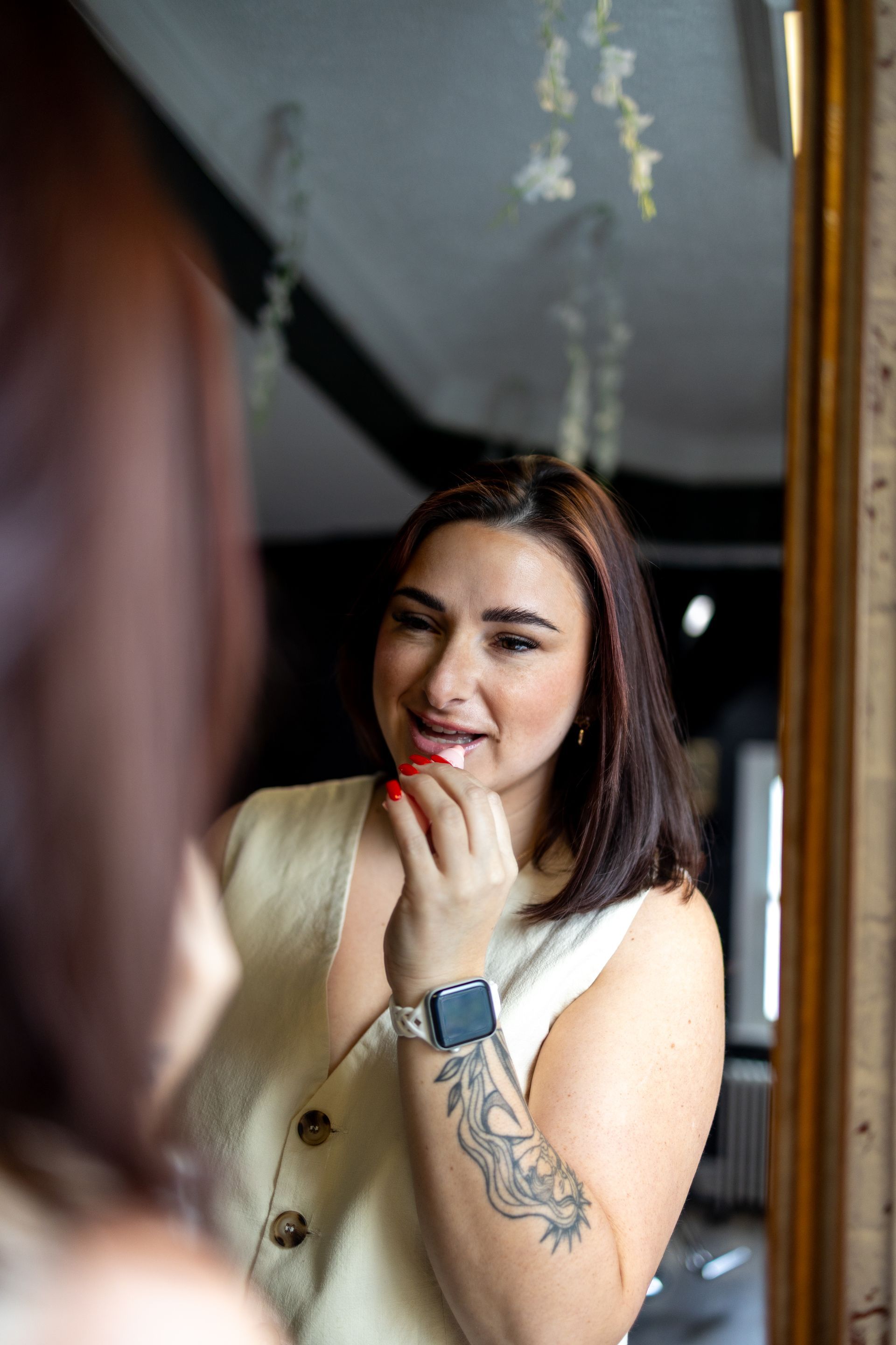 Woman smiling in a mirror, applying lipstick, wearing a sleeveless cream top and a smartwatch.