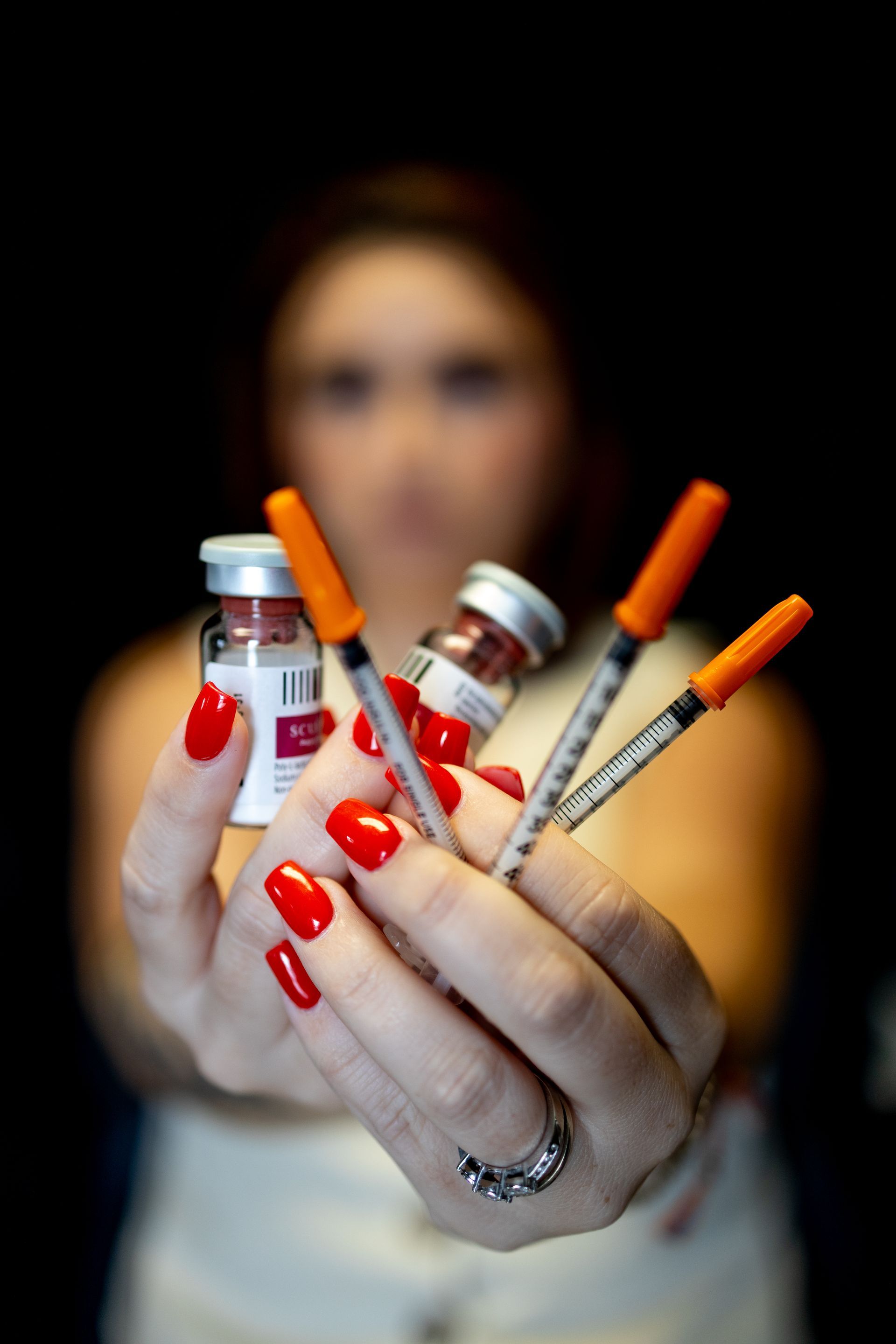Hands with red nails holding three syringes with orange caps and small vials against a dark background