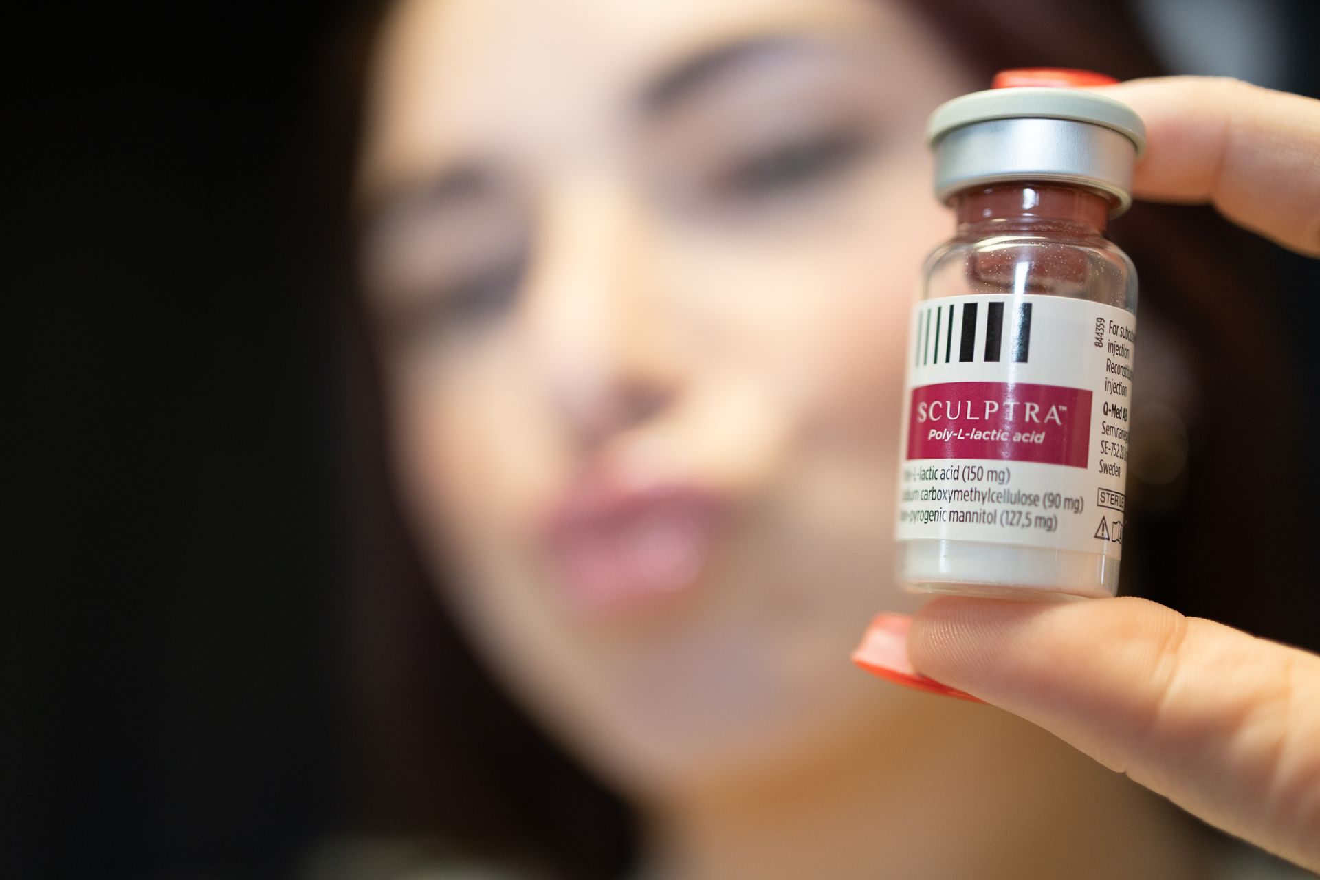 Hand holding a small vaccine vial in focus, with a blurred face in the background